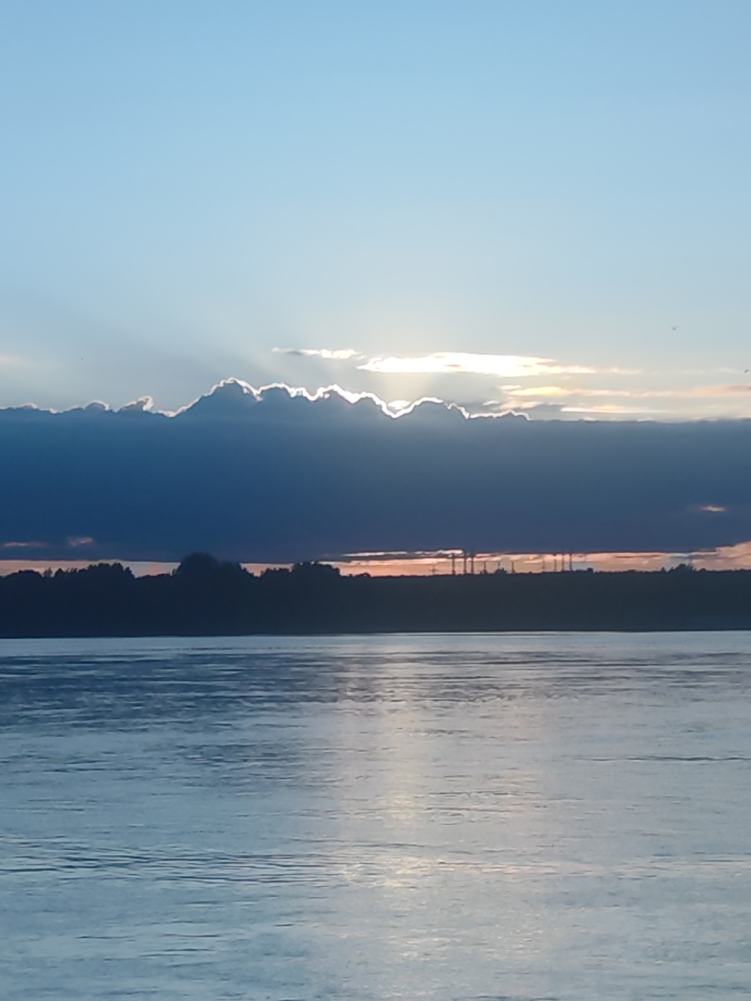 The image presents a tranquil outdoor scene capturing a sunset or sunrise over a vast body of water, likely a river or large lake. The sky is divided into distinct layers: a clear, pale blue at the top, transitioning into a dramatic band of dark, heavy clouds across the mid-section. Bright sunbeams, indicative of the sun being partially obscured by these clouds, radiate outwards, casting a brilliant white glow on the cloud edges and through gaps.
Below the cloud line, a distant landmass stretches across the horizon, appearing as a dark silhouette. Behind this land, a vibrant strip of orange and pink light is visible, characteristic of dawn or dusk. Within this silhouetted landscape, several slender, vertical structures, possibly power line towers or communication masts, can be discerned, suggesting some human infrastructure in the distance. The land itself appears to be covered in trees or dense foliage.
The foreground is filled by the calm surface of the water, which reflects the subdued light from the sky. Gentle ripples create texture and shimmer across the water, giving it a silvery-blue hue. The overall atmosphere of the image is one of quiet contemplation and natural beauty. There are no people visible, nor is there any discernible text within the frame.