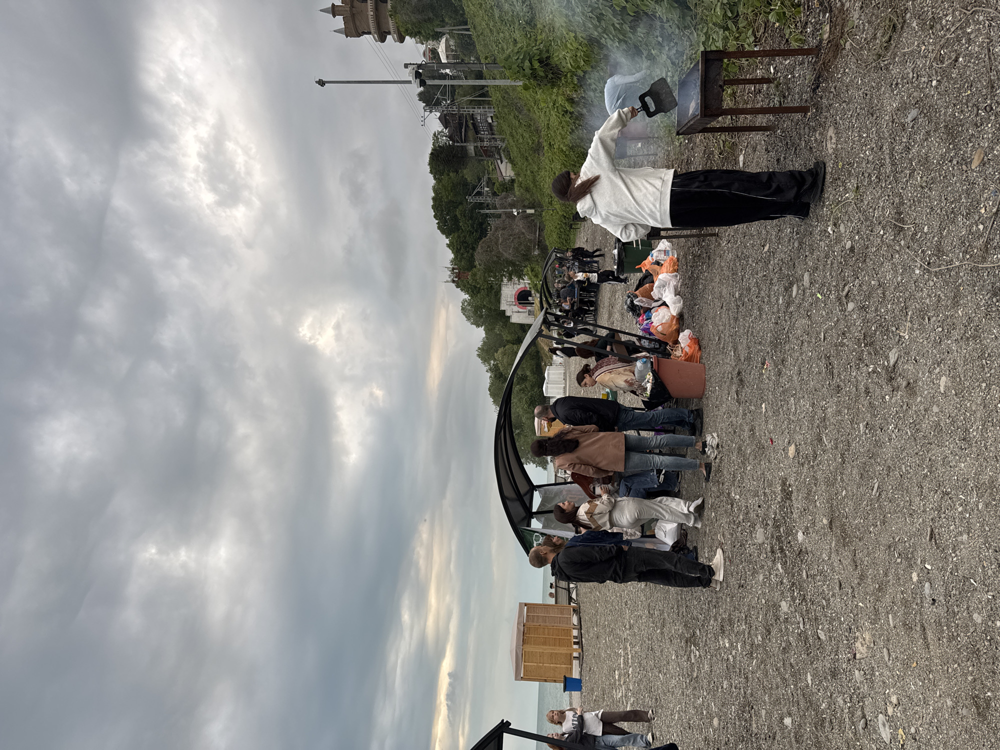 The image depicts a group of people gathered outdoors, likely at a beach or coastal area, given the presence of gravel and a glimpse of water in the distance. Several individuals stand in a line under a canopy structure, while others are seated or positioned nearby. A person stands behind a grill, holding a spatula and tending to what appears to be a barbecue. Bags, containers, and personal items are clustered near the grill, suggesting a picnic or gathering. In the background, buildings and vegetation can be observed, and the cloudy sky indicates overcast weather. Based on the presence of distinctive architecture and landmarks, including the “Sanatorium Ordzhonikidze,” the location can be identified as Sochi, Russia.
