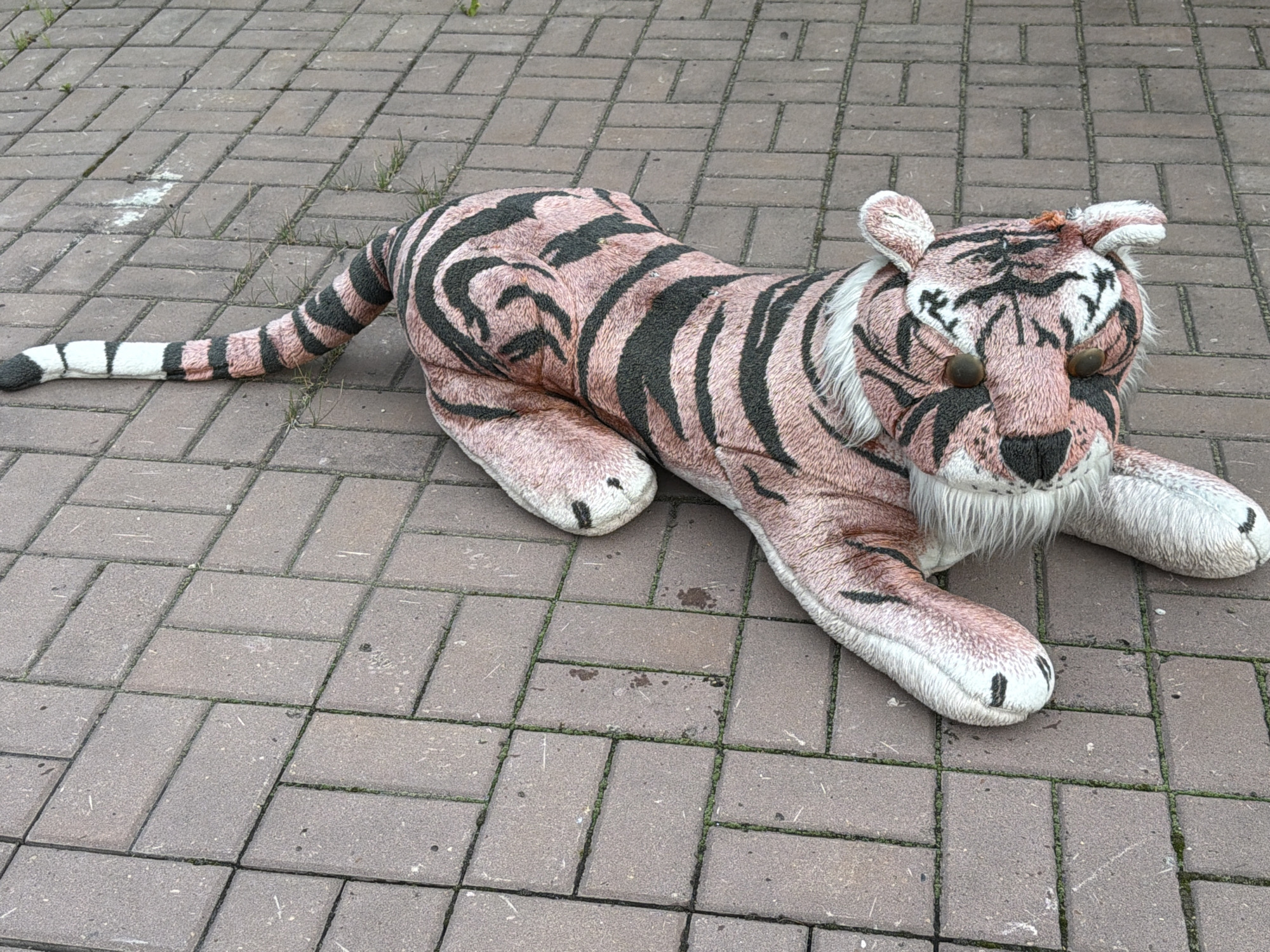 A large, plush tiger stuffed animal lies on a brick-paved ground. The tiger is predominantly pinkish-brown with black stripes, mimicking a real tiger's pattern. It has white fur around its face and chest, and its eyes are round and dark. Its tail extends to the left and ends with white and black rings. The tiger appears to be lying down, with its paws splayed out on the ground. The brick paving is laid in a basket-weave pattern, and small patches of grass are visible in the gaps between the bricks. There are some light-colored smudges on the bricks, possibly chalk or paint. The scene appears to be outdoors, as indicated by the natural light and the presence of grass. There is no visible text in the image. The overall impression is of a toy left outside, possibly a whimsical or slightly melancholic sight. The time of day and weather are not explicitly indicated, but the lighting suggests it is daytime with overcast or diffused sunlight. The location context provided is Каменка, Russia.