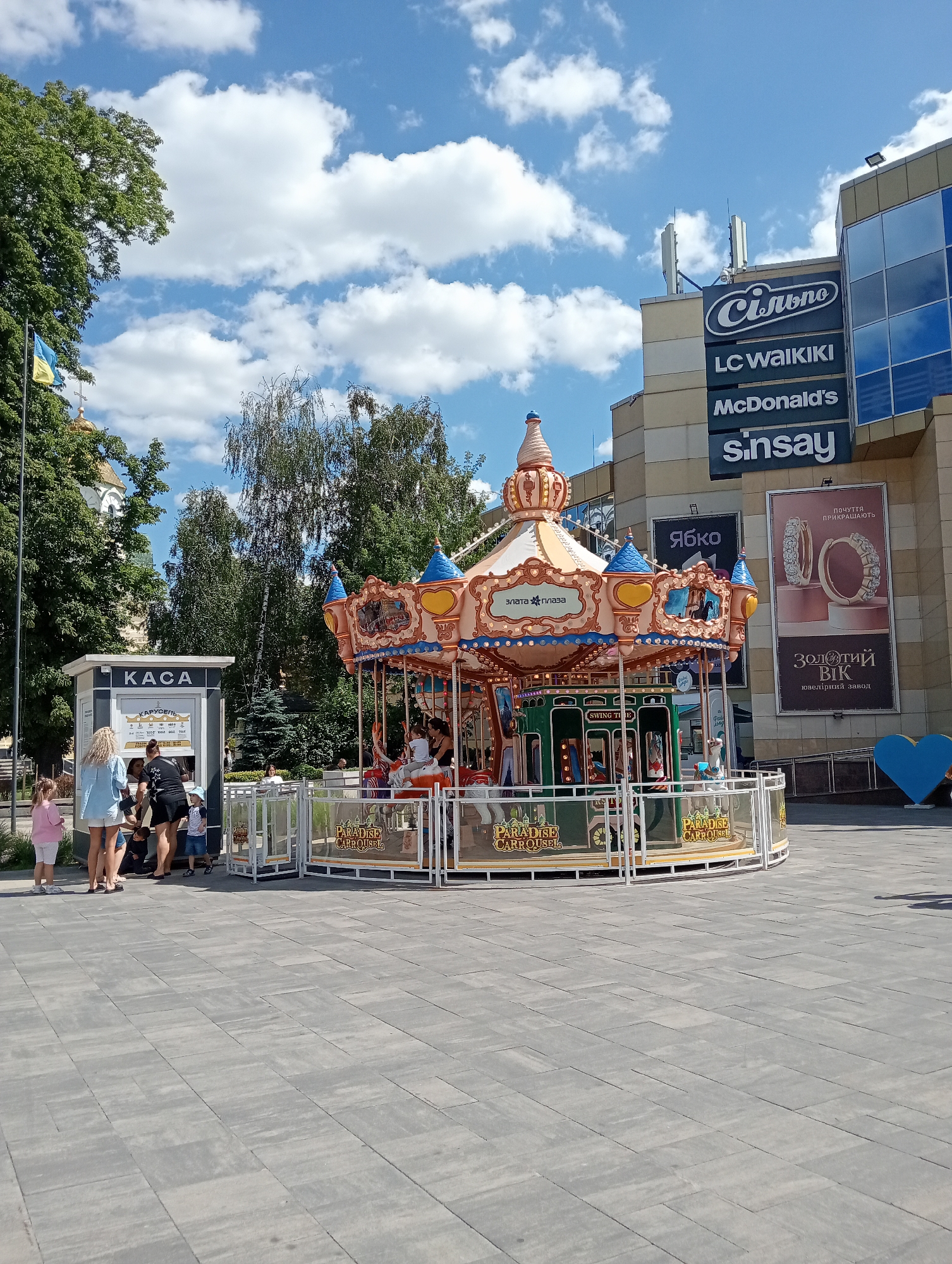 The image depicts a bright, sunny day in the city of Rivne, Ukraine, with a visible Ukrainian flag flying in the background. In the foreground, a colorful carousel, labeled "Paradise Carrousel," stands in an open, paved plaza. Several people, including adults and children, are gathered around the carousel and a nearby ticket booth labeled "KACA." The carousel features various whimsical animal figures and a decorative, castle-like structure at the top. Behind the carousel, a modern building with multiple store signs, including "Silpo," "LC Waikiki," "McDonald's," and "Sinsay," is visible. The architecture suggests a shopping center or commercial complex. The overall scene captures a moment of leisure and recreation in an urban public space.