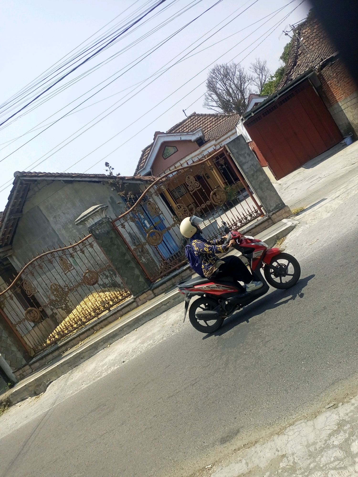 In the city of Ngasem, Indonesia, on a clear, sunny day, a person is riding a red Honda scooter on an asphalt road. The rider is wearing a light-colored helmet, a dark-colored hijab, a patterned long-sleeved shirt, and dark pants. They are positioned on the right side of the road, heading towards the left of the frame. The scooter has a black seat, black wheels with silver accents, and a red and black body.
The background features a residential area with houses and a fence. One house has a grey concrete facade and a tiled roof, with an ornate metal gate in front. Behind the gate, stacked yellow items, possibly agricultural produce, are visible. Another house further back has a tiled roof and a large, dark brown sliding door. The environment is characterized by overhead power lines stretching across the bright blue sky. The road itself is bordered by a raised concrete curb and then the road surface. The perspective of the image is tilted, suggesting it was taken from a moving vehicle or held at an angle.