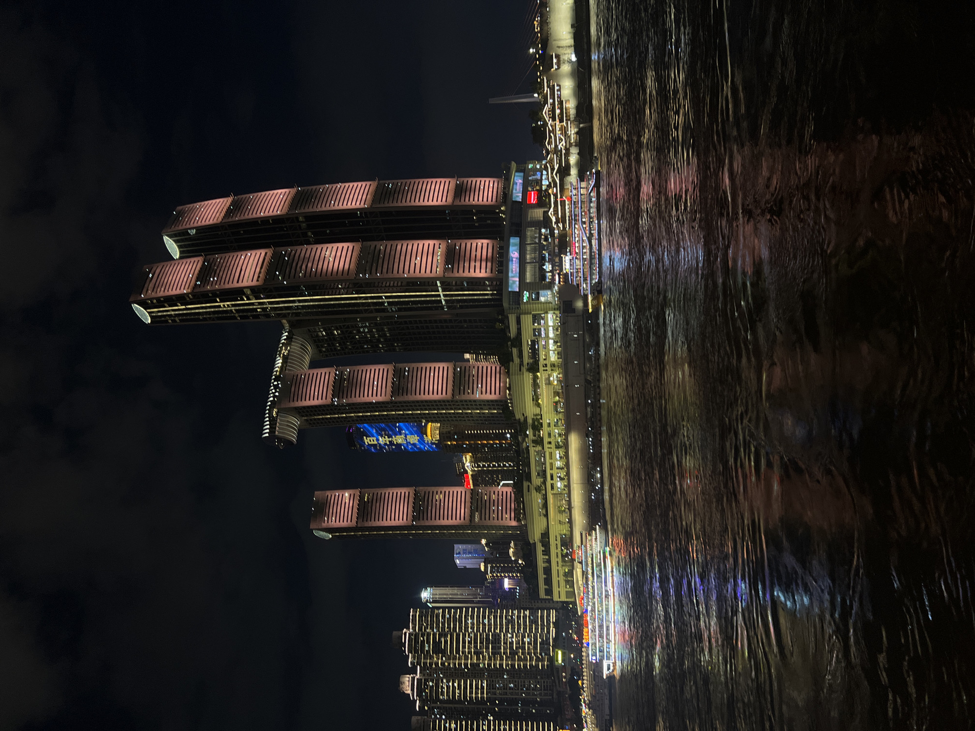 This image depicts a night scene in Nan'an District, China, showcasing a modern cityscape illuminated by artificial lights. In the foreground, a river reflects the colorful lights from the buildings and boats. Two passenger boats are visible on the water, one in the mid-ground and another further to the left. Both boats are brightly lit with multi-colored lights.
The background is dominated by several tall skyscrapers. The most prominent structures are a pair of very tall, slender buildings with reddish-pink lighting. To their left, a cluster of buildings with horizontal lighting creates a dense urban feel. Another tall, slender building with white lighting stands between the reddish-pink towers and the cluster on the left. A distinctive feature of the cityscape is a horizontal structure connecting two of the tallest buildings, resembling a skybridge or elevated walkway. The sky is dark, with some clouds visible. The overall impression is a vibrant and dynamic urban environment at night.