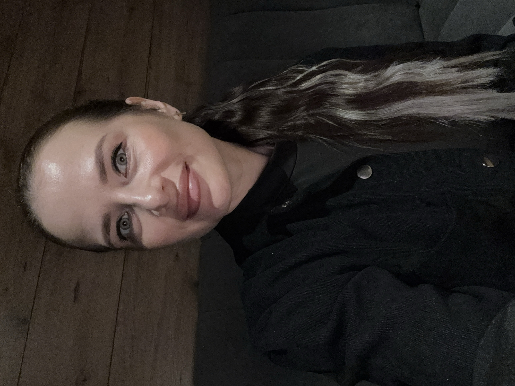 A female subject, with light-colored eyes and a slight smile, gazes directly at the camera in a self-portrait style. Her dark hair is pulled back, featuring a distinct, long streak of lighter grey or silver hair visible on her right side (viewer's left), extending down over her shoulder. She wears a dark, possibly ribbed or corduroy, collared jacket over a dark undergarment. A small, silver-colored cross earring is present in her right ear (viewer's left). The indoor background consists of dark brown horizontal wooden paneling behind her left shoulder and a dark grey upholstered surface, likely a couch or chair, behind her right shoulder and below. The scene is dimly lit. The depicted scene is located in the city of Chaikovsky, Russia.