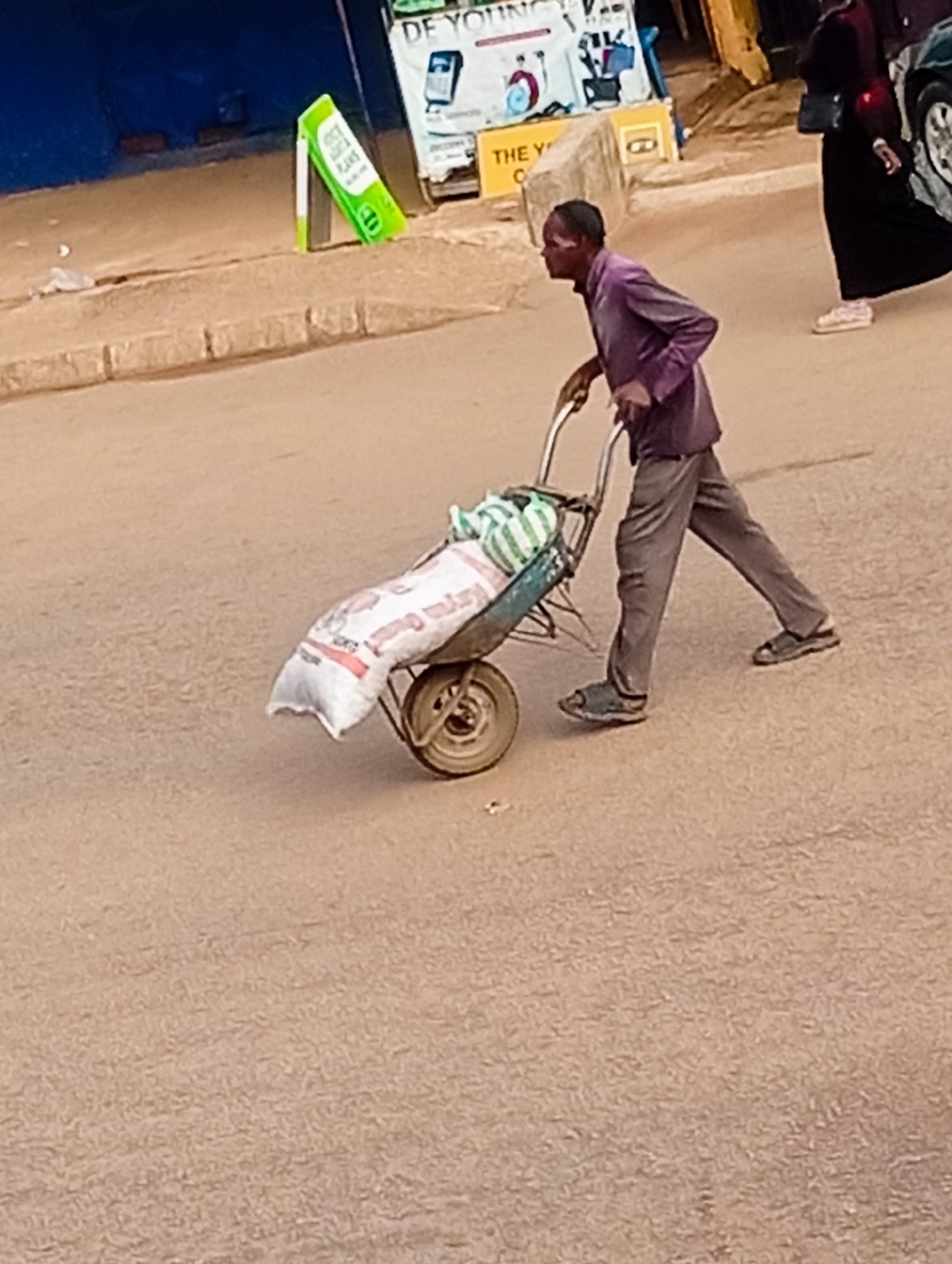 A middle-aged man, wearing a purple shirt and grey trousers, pushes a wheelbarrow laden with two large white sacks and some green and white striped items. He is walking on a paved road in what appears to be a commercial area of Bukuru, Nigeria. Behind him, a woman in a long black dress walks away from the camera. In the background, there are buildings with advertisements, including a green sign that reads "DE YOUNG" and a yellow sign with "THE Y" visible. The road surface is light brown and appears to be dusty.