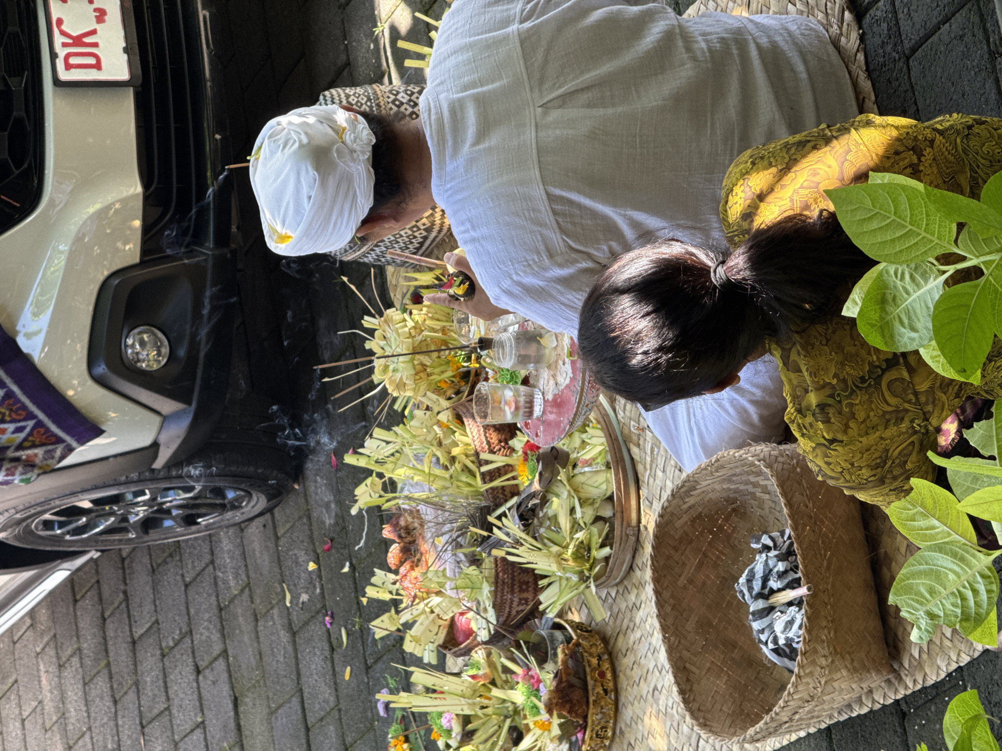 This image depicts a Balinese Hindu blessing ceremony taking place in Ubud District, Bali, Indonesia. An adult male, wearing a traditional white headdress and white clothing, is performing the ceremony. A younger female with black hair pulled into a ponytail is seated beside him. They are positioned on a woven mat alongside a silver car, with incense smoke rising near the front wheel. Several offerings, including baskets filled with flowers, fruit, and woven palm leaves, are arranged in front of them. A woven basket containing cloth sits nearby. The setting is an outdoor paved area, with green plants visible to the right.