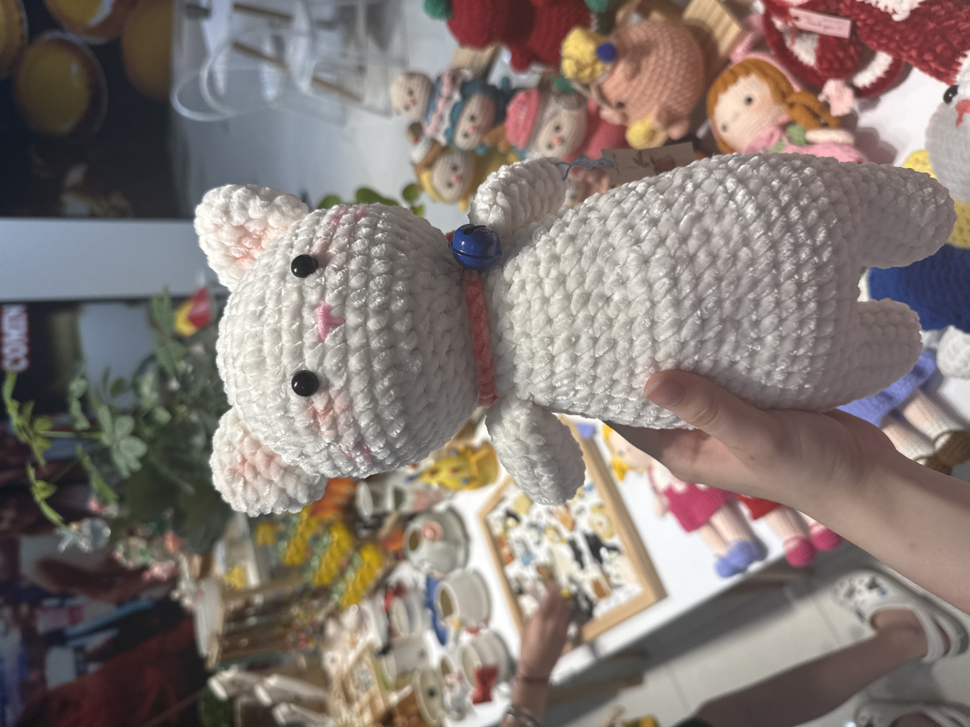 A white, knitted, toy rabbit is being held in a hand. The rabbit has black eyes, pink cheeks, and a blue bell on a red collar. In the background, there are more knitted toys, including dolls and animals. A display case with small objects is visible in the background. Based on the background of the image, it is located at a retail location in Nha Trang Ward, Vietnam.