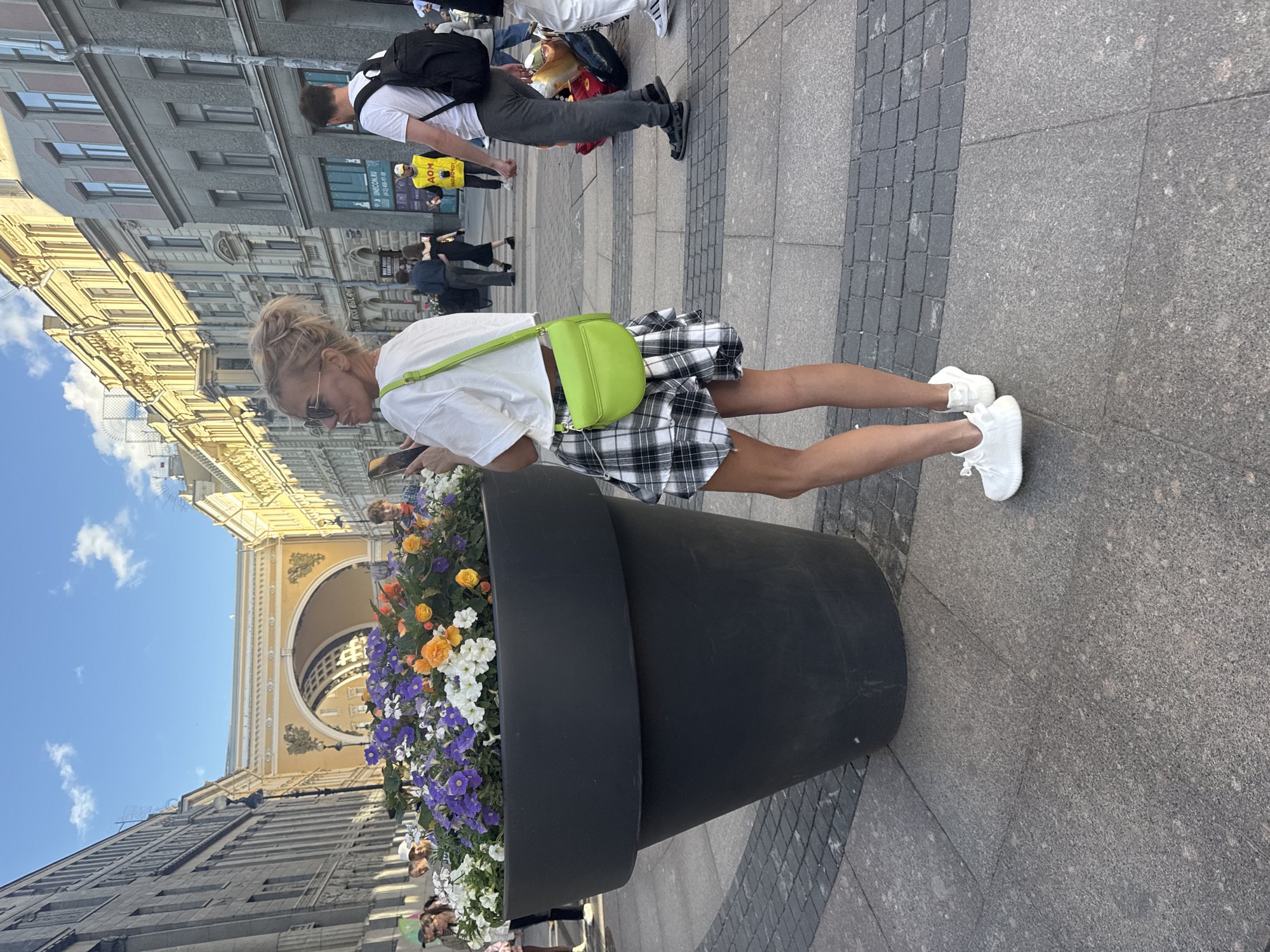 This media file depicts a street scene in Saint Petersburg, Russia. The primary subject is a young woman with blonde hair, wearing sunglasses, a white t-shirt, and a plaid skirt. She is looking down at her phone, which she holds in her right hand. A bright green purse is slung across her body. She is standing next to a very large, black planter filled with colorful flowers, including purple, white, and orange blossoms.
In the background, other people are visible walking along a paved sidewalk. One person wears a yellow vest with visible text. Behind them, classical architecture can be seen, including a large arched building. The sky is blue with some white clouds, suggesting it is daytime and the weather is pleasant. The overall scene appears to be a public area, likely a square or street in a historic part of the city. The woman's posture and engagement with her phone suggest a moment of pause or personal activity within the bustling environment.