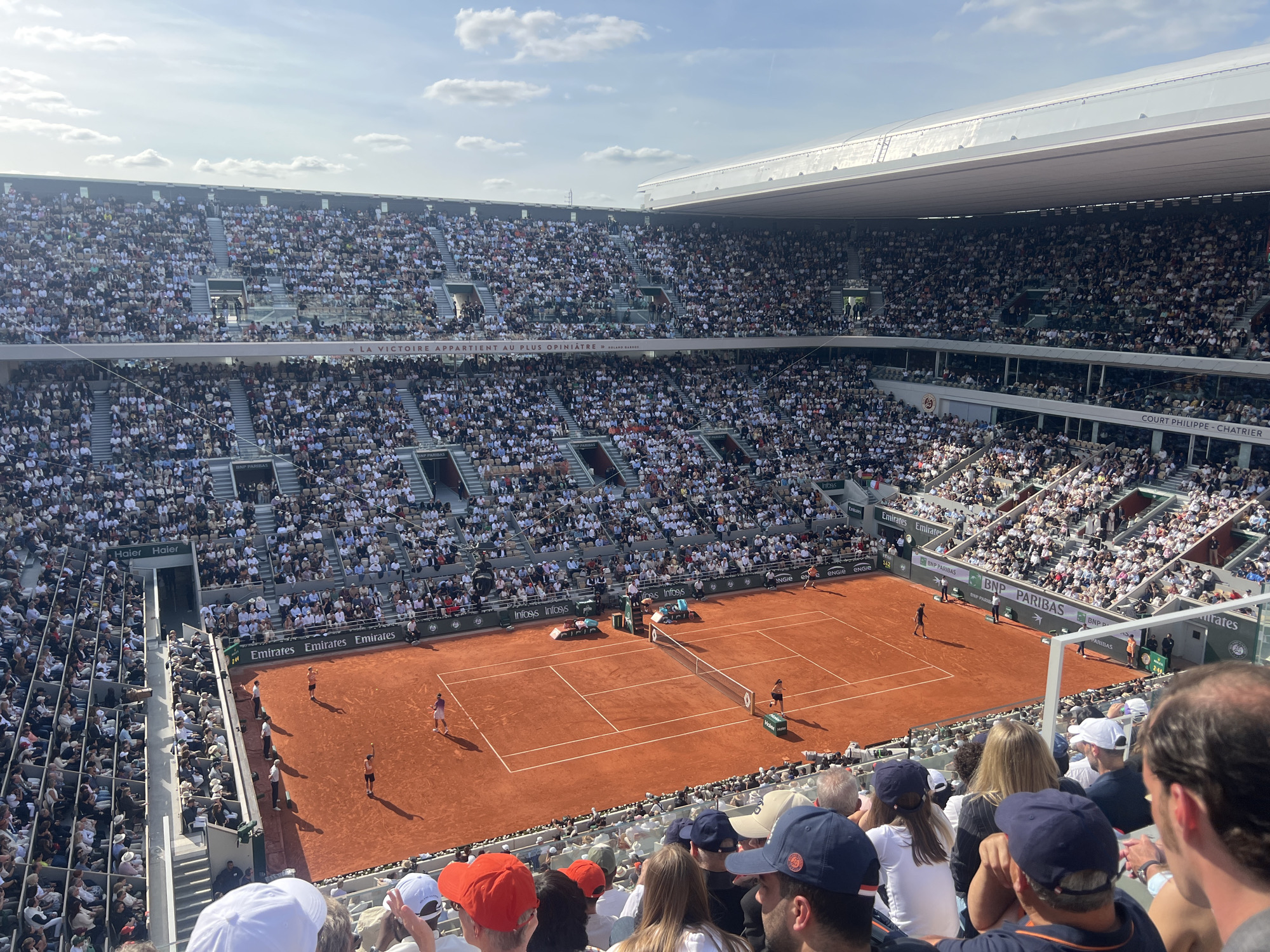 This high-angle, wide shot captures a tennis match in progress at Roland Garros, specifically on Court Philippe Chatrier, located in the 16th arrondissement of Paris, France. The iconic red clay court dominates the lower-middle section of the image, bordered by white lines. Two players are visible on the court: one in purple/dark attire stands near the baseline on the left side, and another in dark attire is closer to the net on the right side. Several officials, including an umpire in the chair and line judges, are positioned around the court.
The stadium is packed with a large crowd of spectators filling multiple tiers of seating, predominantly wearing light-colored, casual clothing. The foreground shows the backs of several audience members, many wearing caps. Advertising hoardings for sponsors such as Emirates, BNP Paribas, Infosys, Haier, and Engie line the court. A banner spanning a section of the upper seating reads, "LA VICTOIRE APPARTIENT AU PLUS OPINIÂTRE" (Victory belongs to the most tenacious), with "ROLAND GARROS" beneath it. The upper portion of the stadium features a visible section of a retractable roof and a clear blue sky with scattered white clouds. The scene is brightly lit by natural sunlight, casting distinct shadows on the court and players.