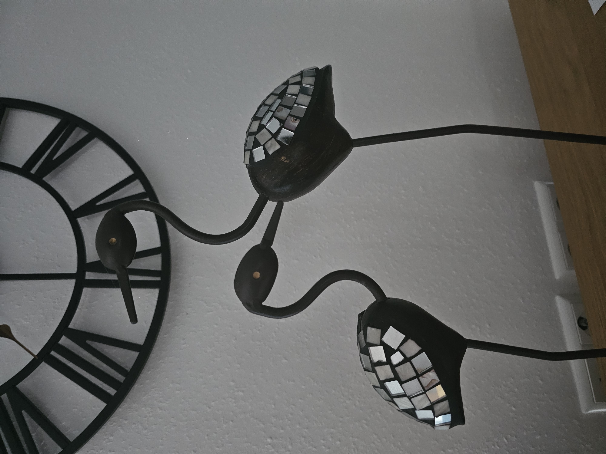 The image shows a close-up, indoor, eye-level shot of two decorative metal flamingo figurines with mosaic tile accents on their bodies, placed on a wooden surface in front of a textured white wall. Above the figurines, a large black wall clock with Roman numerals is partially visible. The clock face is dark, and its hands are also dark. One flamingo is to the left and slightly forward of the other. Both figurines have black, slender necks and heads, with small gold-colored dots for eyes. Their bodies are bulbous and covered in small, square, mirrored mosaic tiles that reflect light. The flamingo on the left has its neck curved down towards its body, while the flamingo on the right has its neck curved upwards, with its head positioned slightly above and to the right of the first flamingo's head, giving the appearance of interaction. The background consists of the white wall and the partially visible clock. The lighting in the scene is diffused, casting soft shadows. The overall aesthetic is decorative and somewhat whimsical.