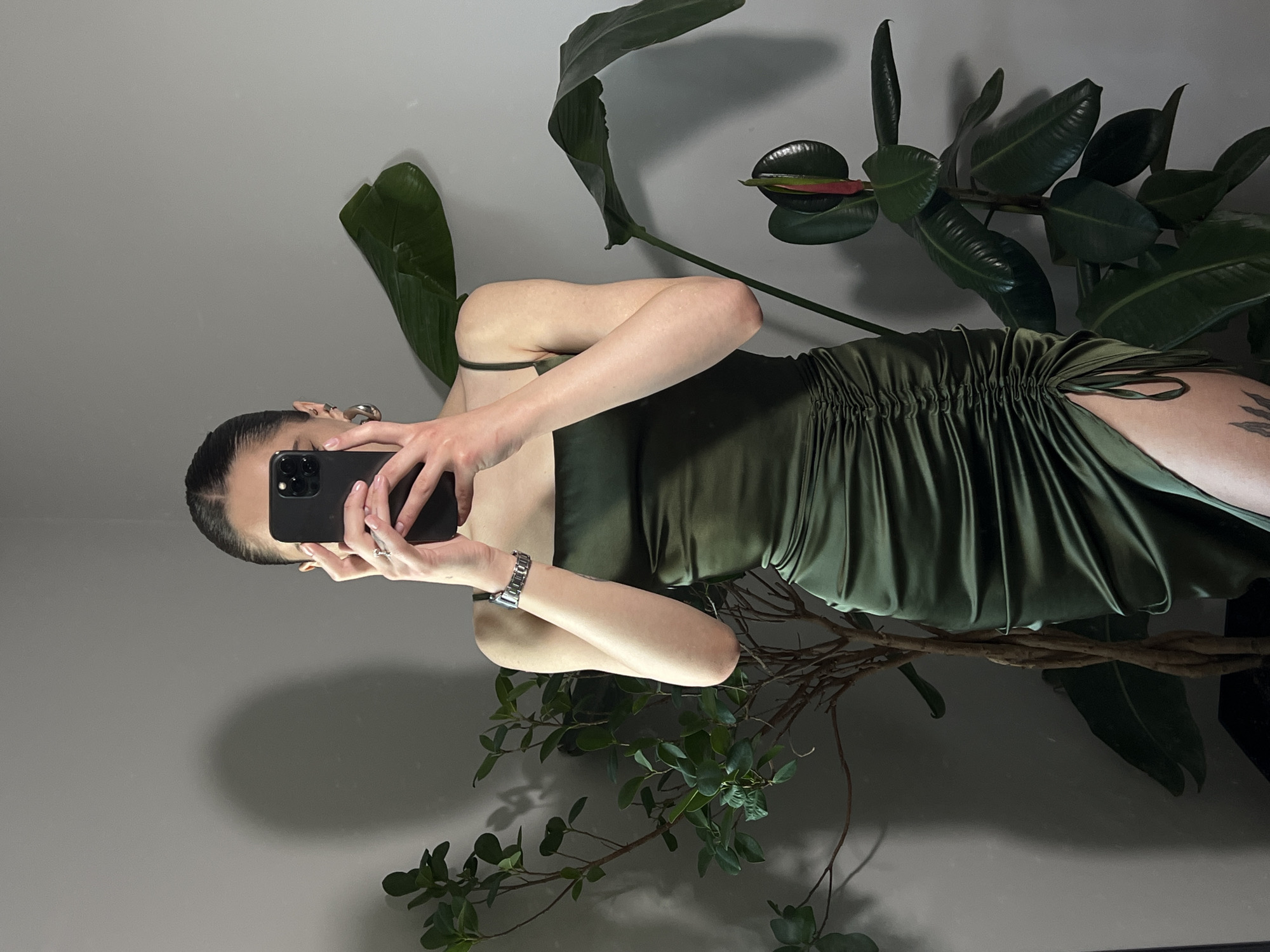 A woman is taking a selfie in a mirror. She is wearing an olive green dress and has her hair pulled back. The mirror reflects a room with plants. The woman is holding a black smartphone. The location is likely indoors, but without additional information, it's not possible to definitively state that it is Ternopil, Ukraine.