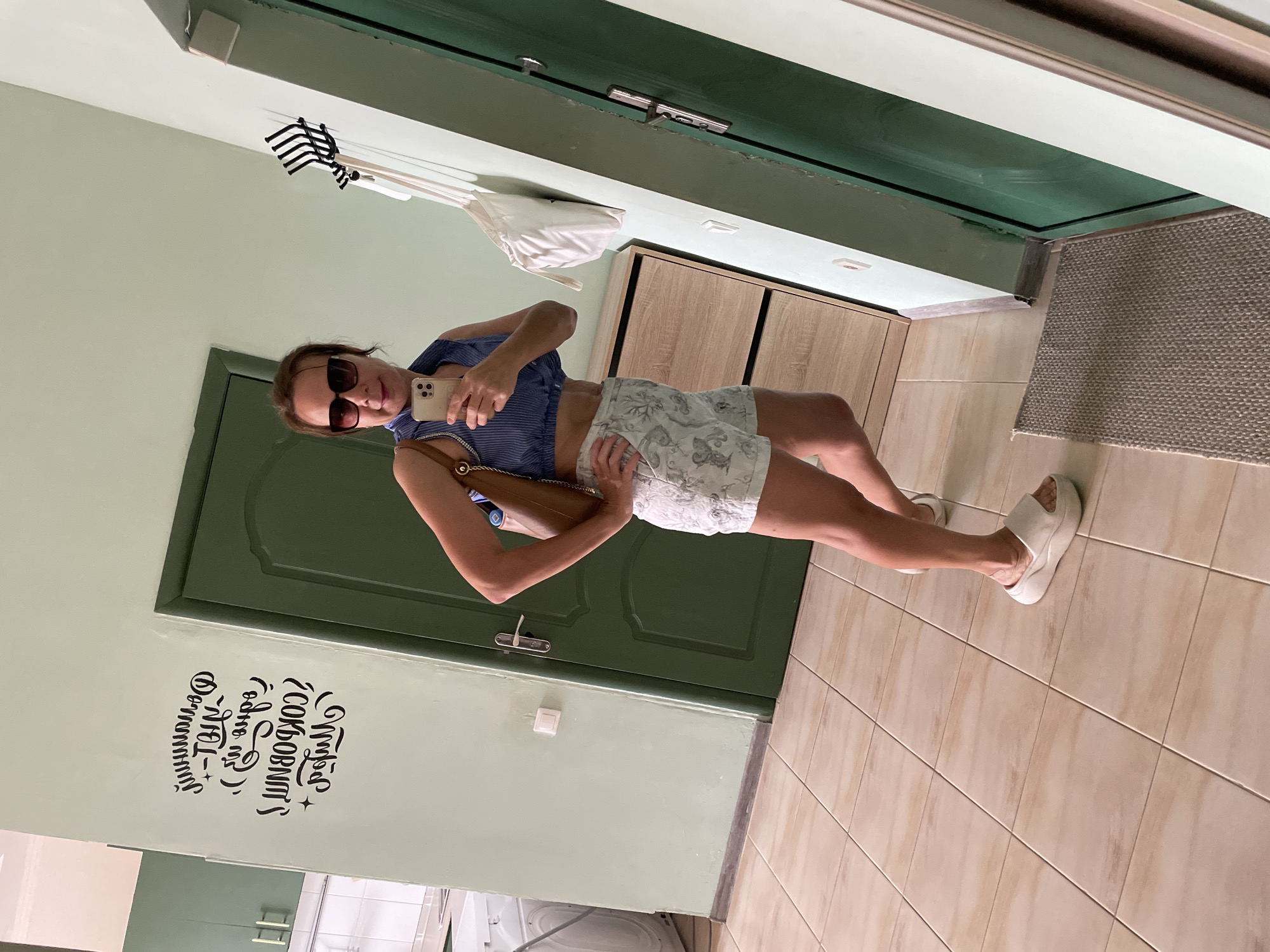 The image shows a woman taking a mirror selfie in what appears to be a hallway or entryway. She is standing in front of a dark green door, with a light green wall to her left and a tiled floor. To her right, there is a beige rug and a wooden cabinet. A white bag and a black rack are hanging on the wall above her.
The woman is wearing a blue and white striped top, white shorts with a floral pattern, sunglasses, and white platform sandals. She is holding a smartphone in her right hand and a brown purse in her left arm. She has brown hair and is looking at the phone.
On the light green wall to her left, there is black text in a stylized font. It appears to be Russian and reads "Не бойся Zоков и Депрессии" which translates to "Don't be afraid of anxiety and depression".
The lighting suggests it is daytime, and the setting appears to be indoors. The overall mood is casual and relaxed.