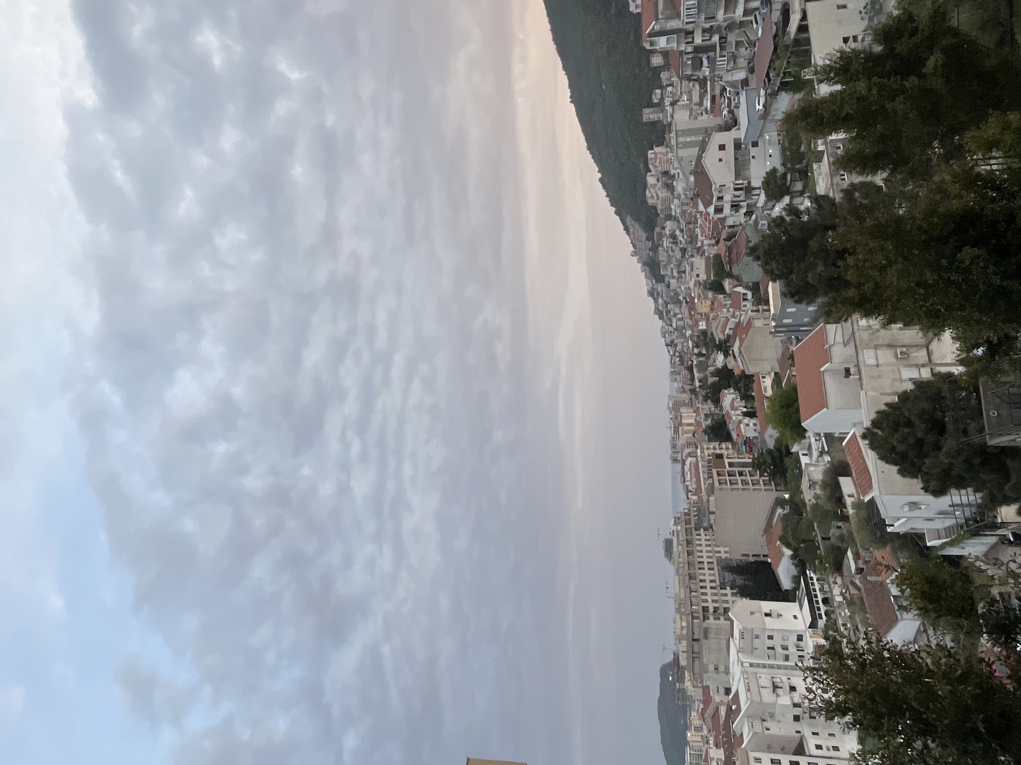 The image displays an elevated view of the coastal city of Budva, Montenegro, appearing to be captured in the late afternoon or early evening. The sky, which occupies the upper half of the frame, is overcast with numerous soft, grey and white stratocumulus clouds. Closer to the horizon, where the sky meets the distant land and sea, there's a faint, hazy glow, suggesting dusk or a low sun.
The right side of the middle ground is dominated by a lush, green mountain slope covered in dense vegetation. Below and to the left of the mountain, the cityscape of Budva unfolds, characterized by a dense arrangement of buildings. These are primarily multi-story structures, mostly residential, with a mix of white, beige, and terracotta-colored facades and roofs. They appear tightly packed together, extending towards the distant sea, which is faintly visible as a hazy line on the horizon. Several dark green trees are interspersed among the buildings, particularly noticeable in the foreground on the right side.
No specific human activity or individuals are visible in the scene. The overall impression is that of a bustling coastal town nestled against a mountain backdrop, under a serene, muted sky. There is no visible text in the image.