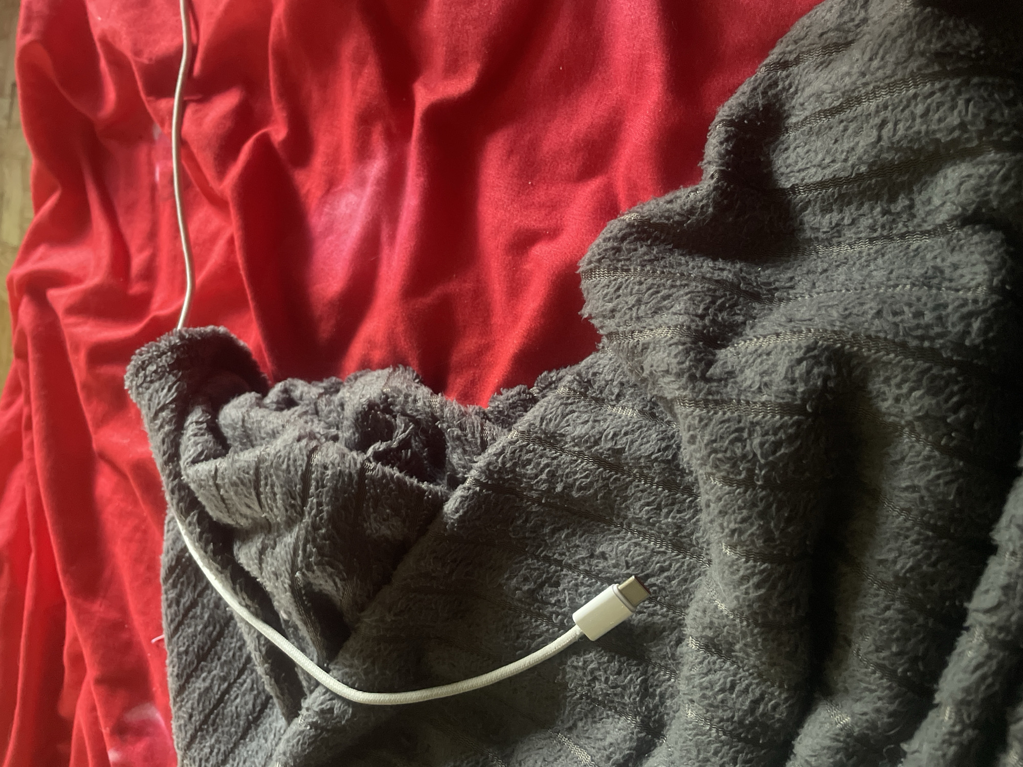 The image shows a close-up of a crumpled gray fleece blanket with a white USB-C cable resting on it, partially on a bright red fabric background. The blanket has a textured, fuzzy surface with thin, vertical stripes. The red fabric is creased and appears to be a sheet or similar material. The scene is indoors, with the lighting suggesting an ambient setting. There are no people visible in the image. The overall impression is one of casual disarray. The image location in Линёвка, Russia cannot be determined from the visual information provided.