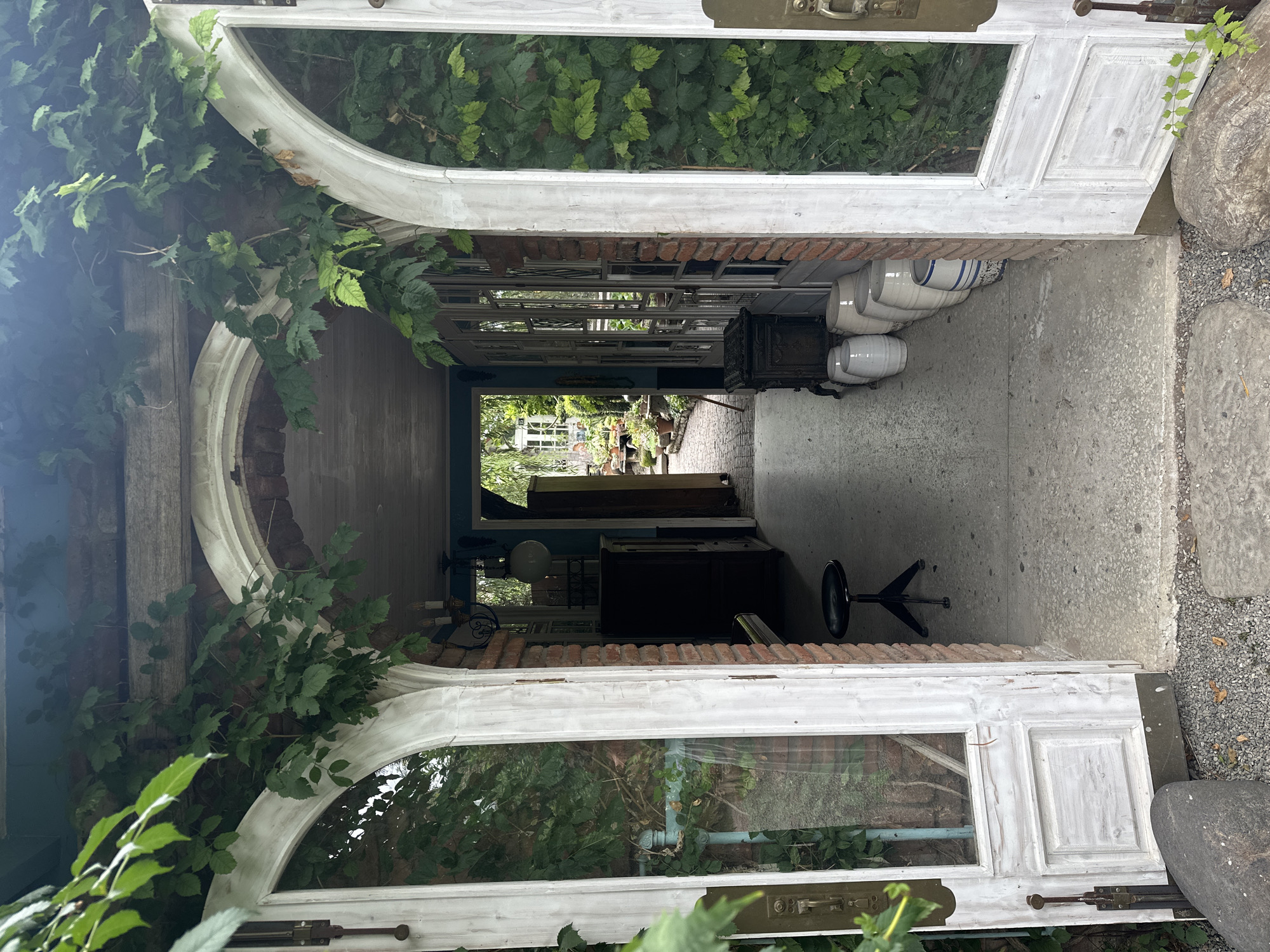 This is a wide-angle, eye-level outdoor shot of an arched doorway opening into a walkway or alley. The doorway features two large, white, rustic French doors, both ajar. Lush green vines are growing over the arched structure and the brickwork to the right of the doorway, partially obscuring it.
The walkway beyond the doors is paved with concrete and gravel. To the left, inside the doorway, a dark, tall cabinet or hutch is visible, and a black, swivel stool with a metal base stands in front of it. To the right of the walkway, a small, dark, cast-iron stove sits on the concrete. Lined up against the brick wall to the right are several large, white ceramic pots or crocks, some with blue stripes.
The walkway extends into the distance, where more greenery and what appears to be a garden with potted plants and structures can be seen. The overall impression is one of an overgrown, charming, and slightly whimsical outdoor space, possibly a courtyard or garden entrance. The light suggests it is daytime, with some areas in shadow. The location is not identifiable from the image itself.
