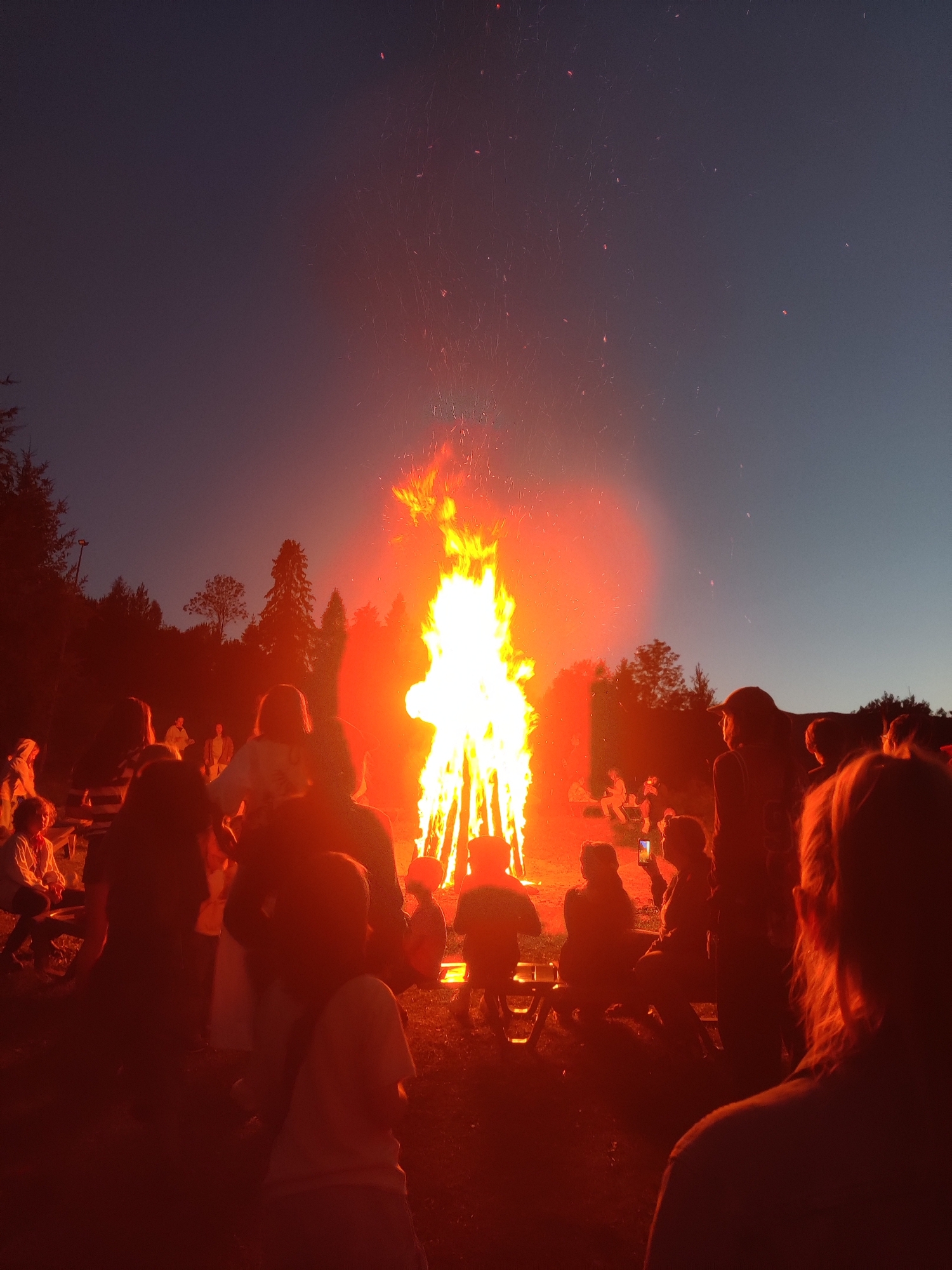 A group of people are gathered around a large bonfire at dusk in what appears to be a rural outdoor setting, possibly in Izky, Ukraine. The fire is the dominant light source, casting a warm orange glow on the silhouetted figures. Trees and a hillside form the background, against a darkening sky with a hint of twilight. Some individuals are sitting on benches or the ground, while others are standing, observing the flames. A few sparks are visible rising from the fire into the air. The overall scene suggests a social gathering or event centered around the bonfire.