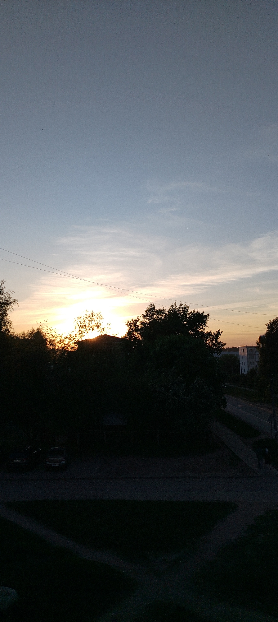 The image shows a sunset view from a high vantage point, potentially a building window in Chaikovsky, Russia. The sky transitions from light blue to orange-yellow near the horizon. Silhouetted trees and possibly a building obscure the direct view of the sun. Below the trees, a road and a sidewalk are visible, with what appears to be a person walking on the sidewalk. Parked cars are seen on the left side. The foreground area is dark and appears to be a grassy or paved surface. Power lines stretch across the sky. In the distance, a multi-story building can be seen.