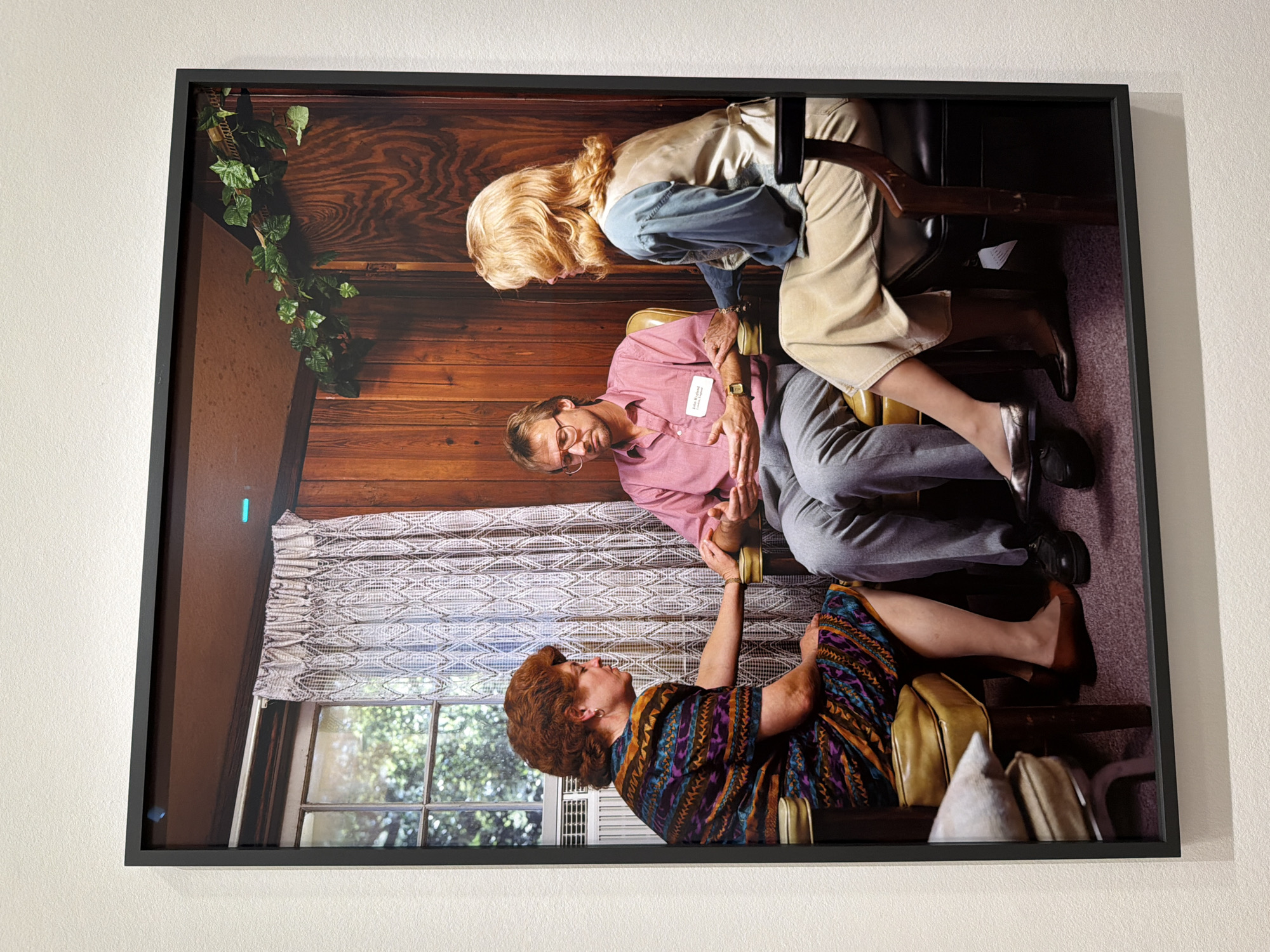 This photograph, rotated 90 degrees clockwise, captures an intimate and complex interaction between three individuals within an indoor setting.
The scene unfolds in a room characterized by warm wood-paneled walls and a window with a patterned fabric curtain, revealing lush green foliage outside, indicating daytime. Artificial ivy is draped in the upper left corner, and an air conditioning unit is visible below the window. The furniture includes at least three chairs, two of which are upholstered in a light tan color.
At the center of the image, a man with glasses, wearing a pink collared shirt and gray pants, is seated. A name tag on his shirt reads "John R. W_____" and "Licensed Counsel_____", identifying him as a licensed professional. He wears a gold watch on his left wrist, and his hands are clasped. Above him (in the rotated view), a blonde woman in a light-colored vest, blue shirt, and light pants leans over, seemingly reaching towards the man's hands or lap. Below the man, a woman with reddish-brown curly hair, dressed in a brightly patterned dress (purples, blues, yellows), sits on another chair, gently holding the man's foot.
The expressions of the visible faces are neutral and contemplative, suggesting a serious or therapeutic discussion or encounter. The unique positioning of the subjects creates a sense of focused engagement and emotional depth. The interior décor, with its wood paneling and furniture style, evokes a somewhat dated aesthetic.
This artwork is observed in Paris, France.