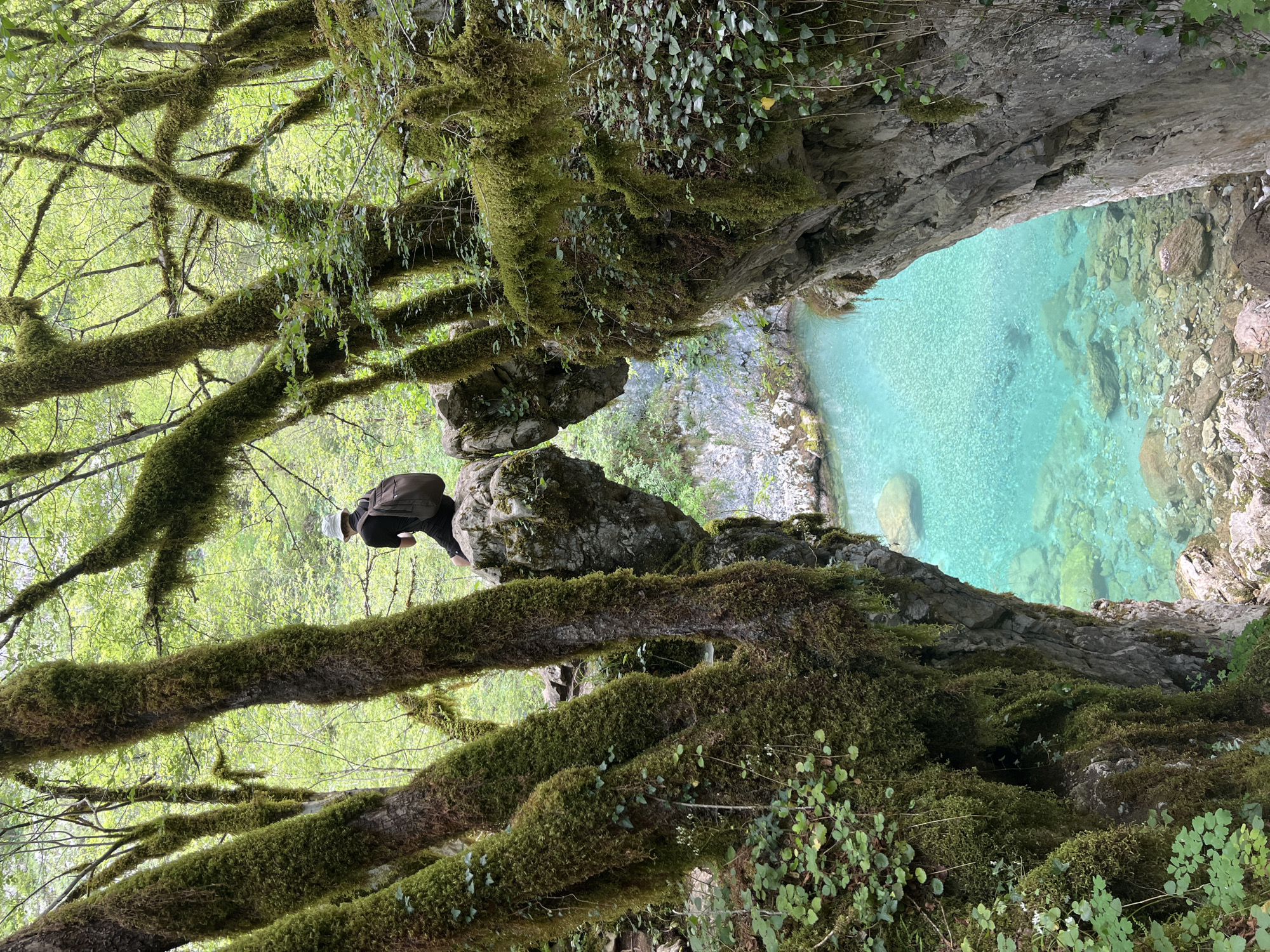A hiker with a backpack, wearing a white hat and dark clothing, is perched on a moss-covered rocky outcrop. Below him, a clear turquoise river flows through a rocky gorge. The scene is framed by moss-covered tree branches and rocks. The location appears to be outdoors and the time of day is daytime. Based on the prompt, the location is the city of Lazba, Montenegro.