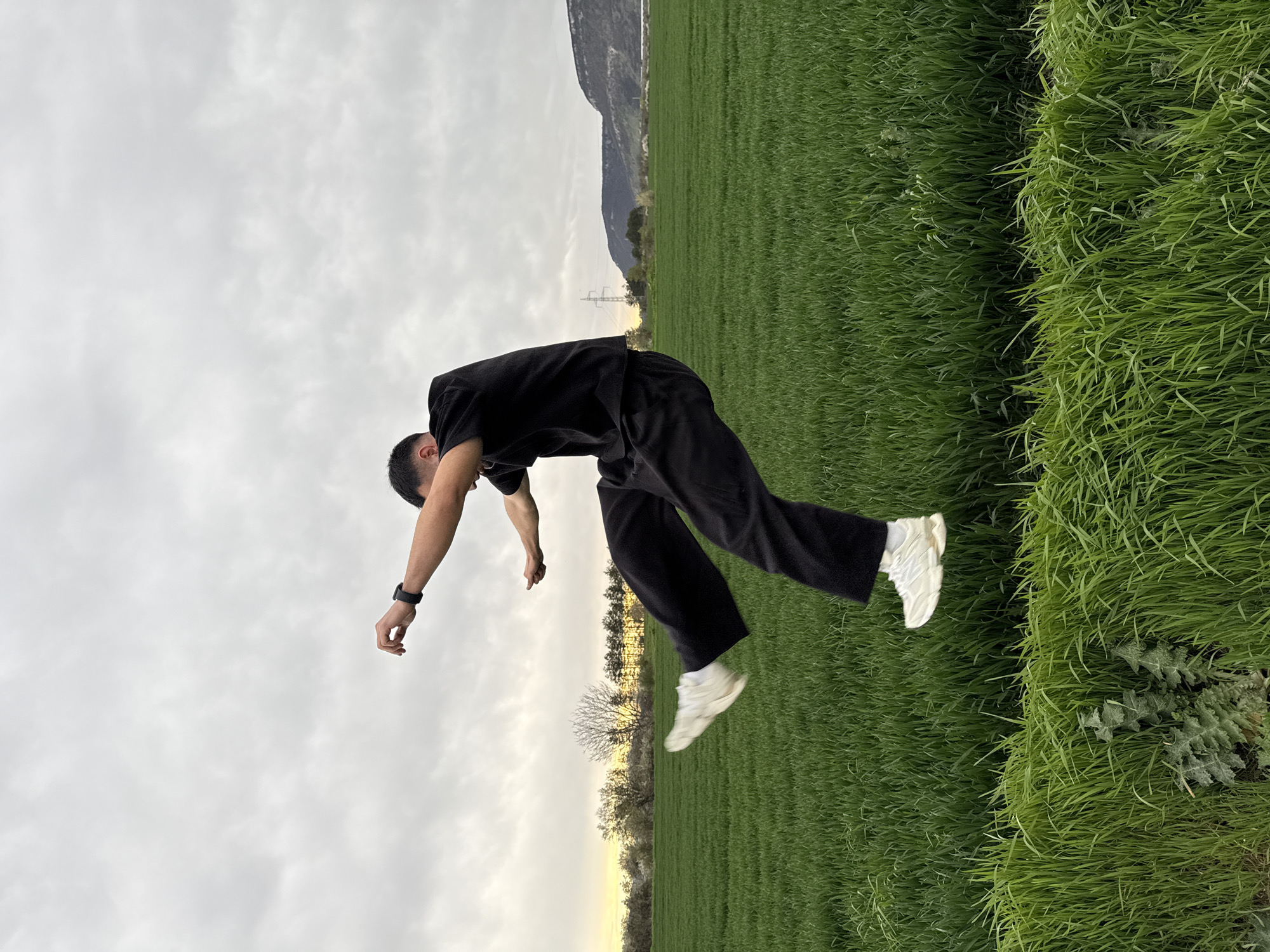 A man is suspended horizontally in mid-air, parallel to a field of green grass. He is wearing a black t-shirt, black pants, and white sneakers. His body is slightly arched backwards, with his arms extended downwards. He is also wearing a black watch on his left wrist. The background consists of a green field, distant hills, a power line, trees, and a cloudy sky. The image has been rotated counterclockwise. The exact location in Shumen, Bulgaria, cannot be determined from the image.