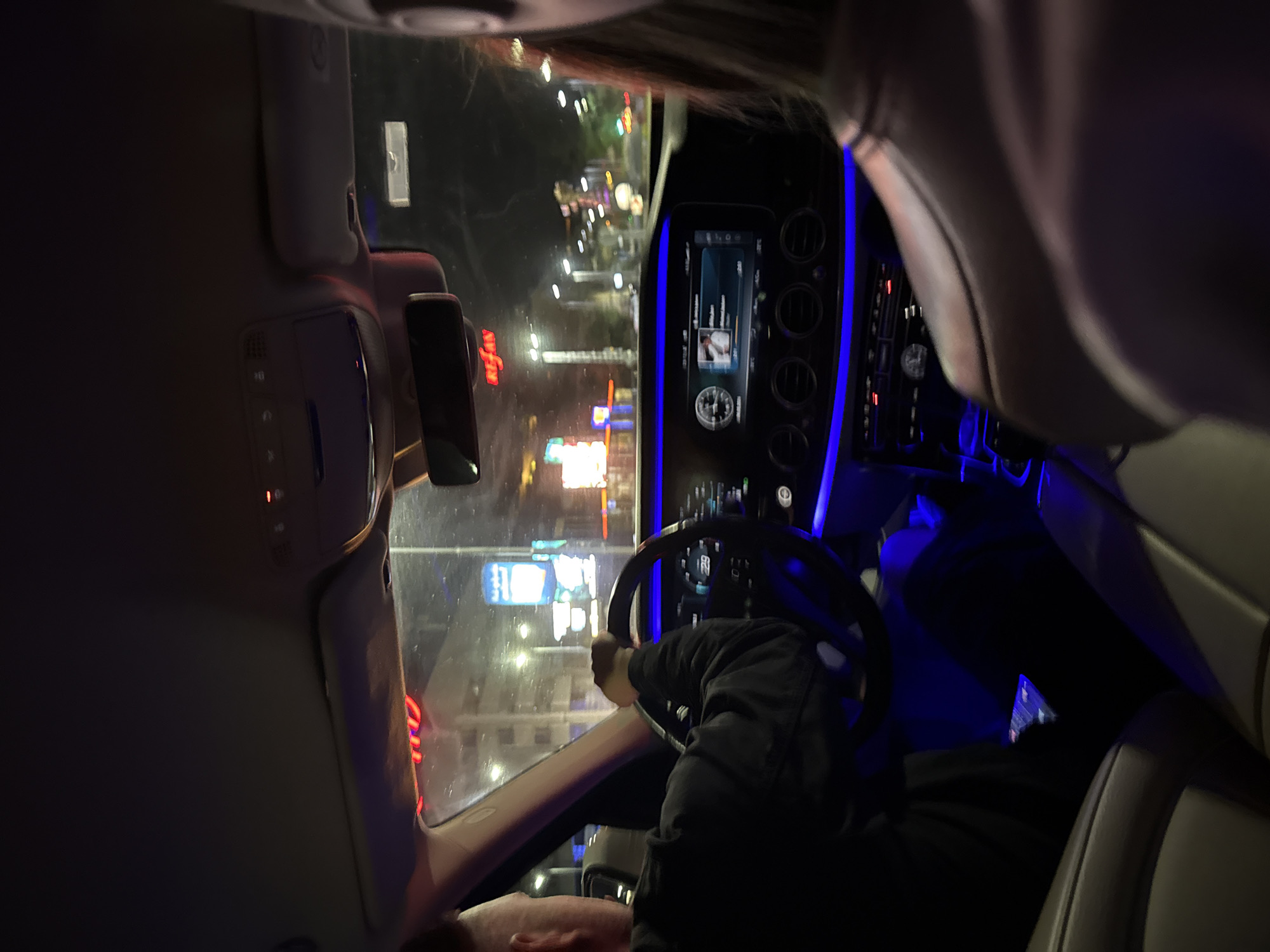The image depicts the interior of a car at night. The driver is holding the steering wheel with one hand, with a person visible in the passenger seat. Outside the car's windshield, the city lights of Plovdiv, Bulgaria are visible. The car's dashboard is illuminated, and the infotainment screen is displaying an image. The ambient lighting inside the car is blue.