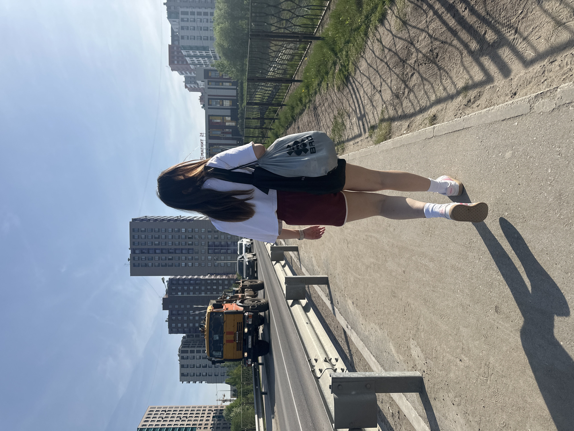 The image depicts a scene in Perm, Russia, with a pedestrian standing on a bridge. The bridge appears to span a road with heavy machinery, possibly construction equipment. The pedestrian is wearing a white top and dark shorts, with long brown hair. They are carrying a gray bag with black straps. High-rise buildings are visible in the background, suggesting an urban environment. The day appears sunny, based on the shadows and the sky. The bridge has metal railings.
