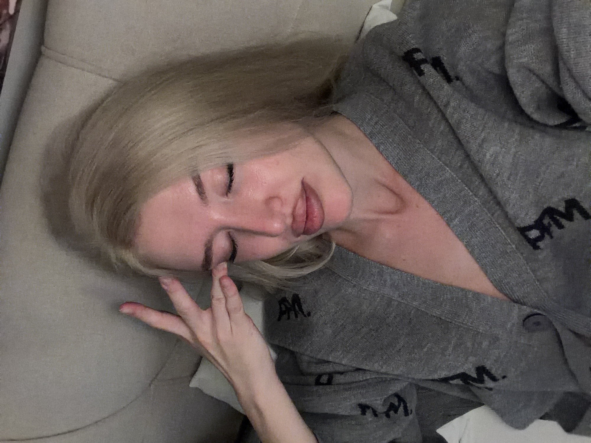 A blonde female is observed indoors in Belgrade, Serbia, positioned against a light-colored, upholstered background, possibly a headboard or sofa. Her eyes are closed, and her lips are slightly parted, exhibiting a glossy appearance. Her right hand is raised, with fingers splayed, and the index finger and thumb are pressed against her right temple area. She is wearing a grey knit cardigan adorned with dark letters, possibly "PM" or "PIM", and a single dark button is visible on the garment. A white fabric, resembling a pillow or sheet, is visible behind her left shoulder. The scene is lit with soft, direct lighting, creating a visible shadow on the background upholstery.