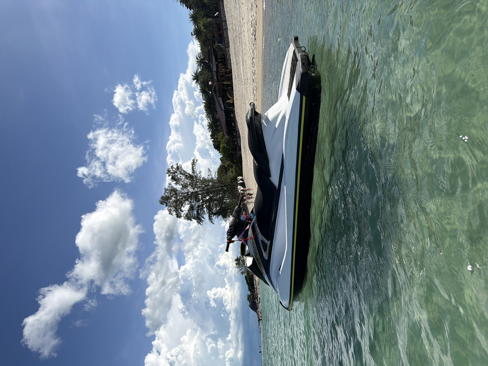 A jet ski is moored in clear, turquoise water near a sandy beach. The jet ski is white with black accents and a yellow stripe along its side. In the background, palm trees line the beach, and a few buildings with thatched roofs are visible. The sky is a brilliant blue, dotted with fluffy white clouds. People are visible on the beach and in the water in the distance, some on other watercraft. The scene appears to be a tropical resort destination.