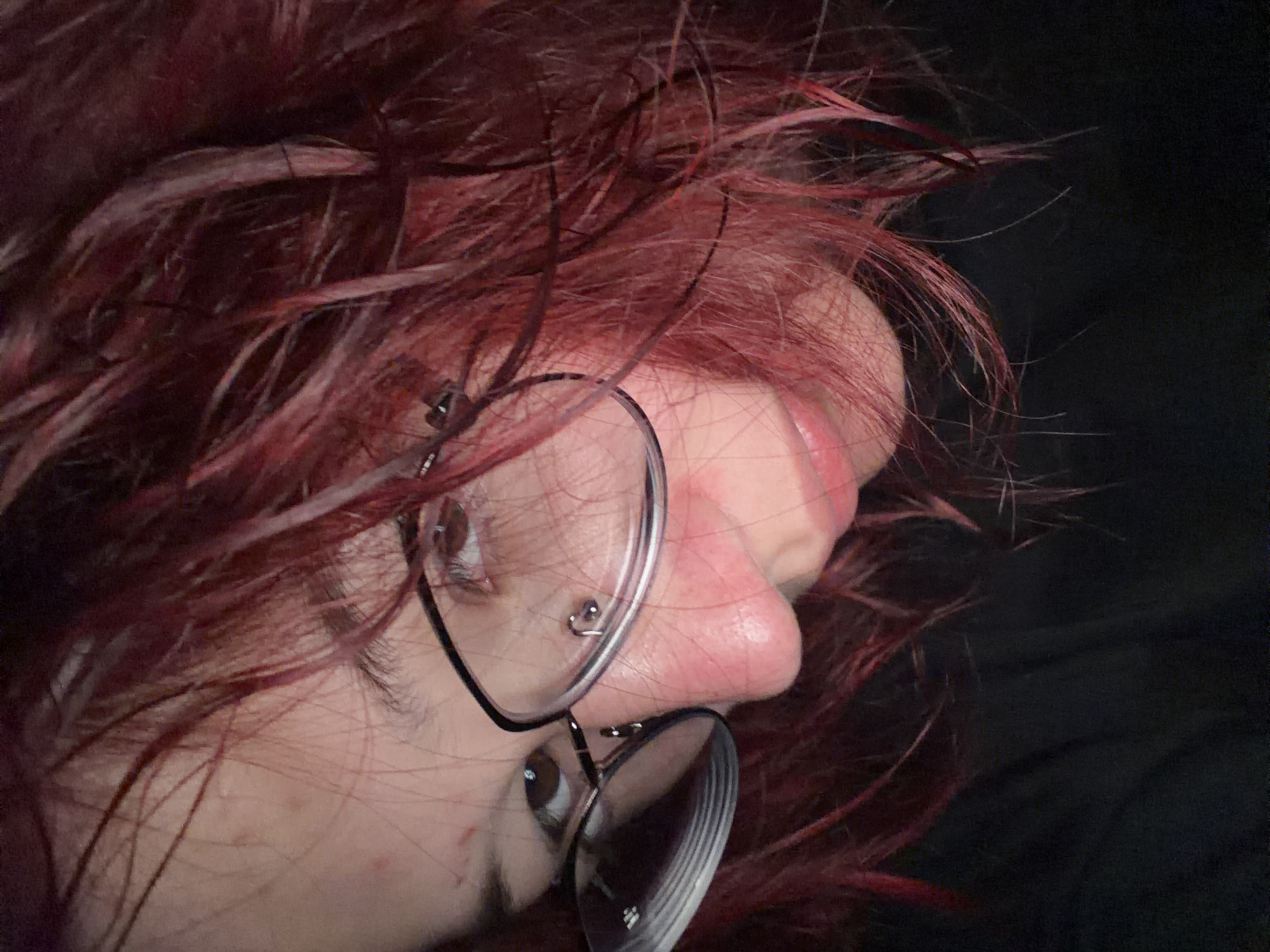 A close-up, low-angle shot shows the face of a person with dark red, messy hair, wearing black-rimmed glasses and a nose piercing. The person's skin appears pale, with a few small blemishes visible on the forehead. The lighting is dim, casting shadows and highlighting the texture of the hair and skin. The person is looking downwards and slightly to the right. The background is dark and out of focus, suggesting the shot was taken indoors in low light conditions. There is no discernible location in Chelyabinsk, Russia.