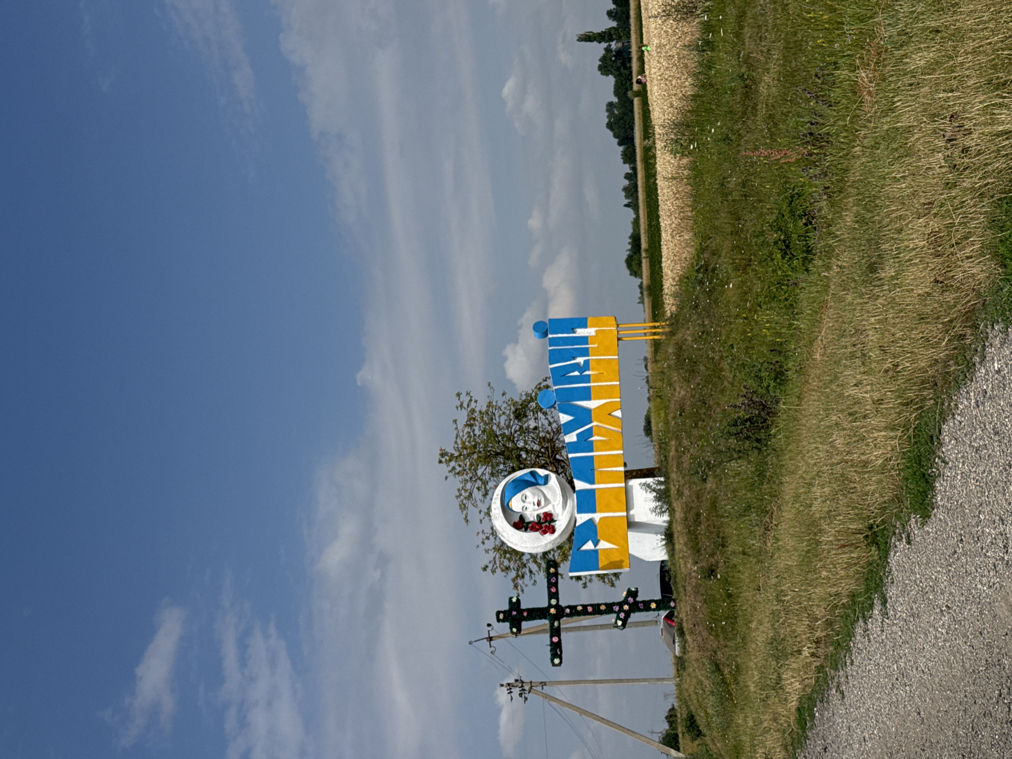 The image shows a roadside monument in Vilkhivtsi, Ukraine, under a blue sky with scattered clouds. The monument features a large white circular emblem with a painted portrait of a woman wearing a blue headscarf and holding red roses. Below the emblem, large blue and yellow letters spell out what appears to be the name of the town. The monument is supported by a white base and yellow metal legs. To the left of the monument is a dark green, cross-shaped structure decorated with colorful flowers. In the background, a field of golden wheat stretches towards a line of trees. A power line pole is visible to the far left, and a gravel road runs along the bottom edge of the frame. Two figures, likely women, are visible in the distance in the wheat field.