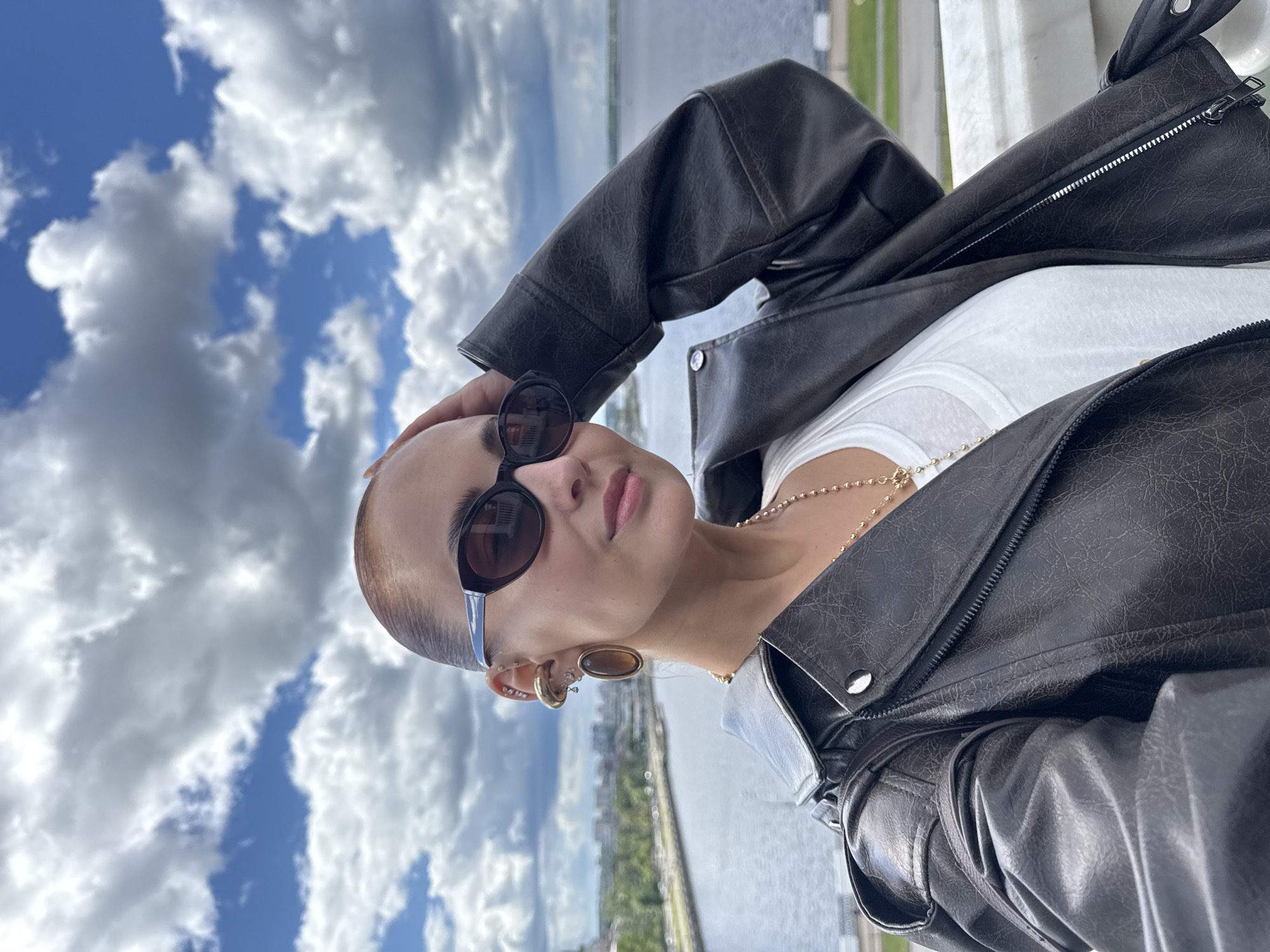 A medium, vertically-oriented shot depicts a woman outside on a bright day, with a backdrop of cloudy sky, river, and city buildings. She is wearing sunglasses, a gold necklace, and a dark leather jacket over a white shirt. The woman has short, light brown hair and is holding her head with her left hand. She also has multiple earrings on her left ear. In the background, a wide river stretches towards a cityscape that includes green spaces and tall buildings. The clouds are prominent, with a mix of white and gray formations against a blue sky. Based on the visual elements, it is not possible to confirm the location as Izhevsk, Russia, without additional contextual information.