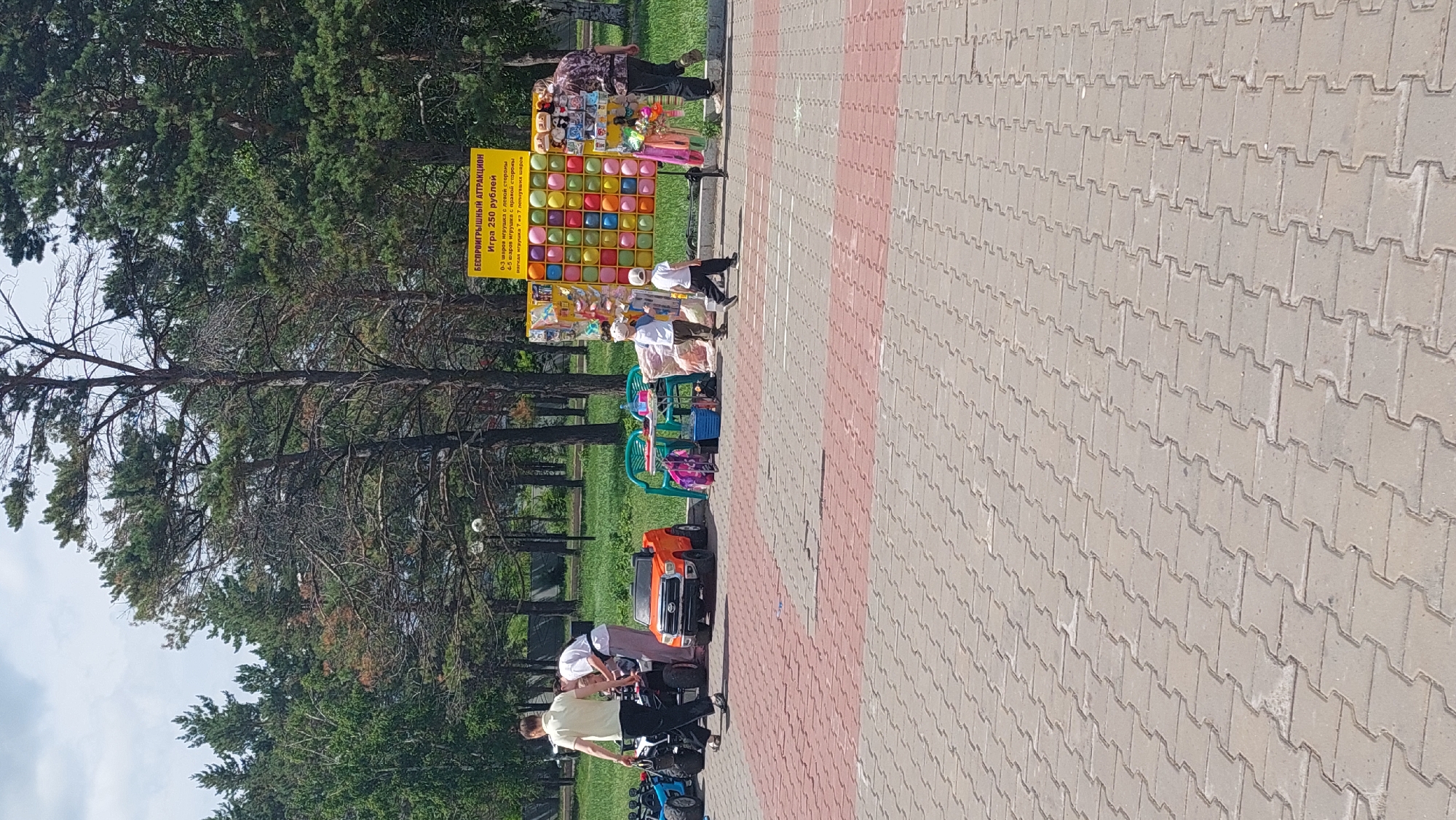 An outdoor scene depicts a section of a paved walkway in Komsomolsk-on-Amur, Russia. A yellow banner advertises a "Guaranteed Prize Attraction" for 250 rubles, featuring a wall of colorful balloons. Two young children, both wearing white caps, stand near the balloon wall, with one child appearing to be walking away. To the left, a person in a light-colored shirt and dark pants walks towards a small orange toy car, beside which are two other toy vehicles. A woman in a white shirt and pink pants is seen tending to something near the toy cars. In the background, numerous tall trees line the area, and the sky is partly cloudy.