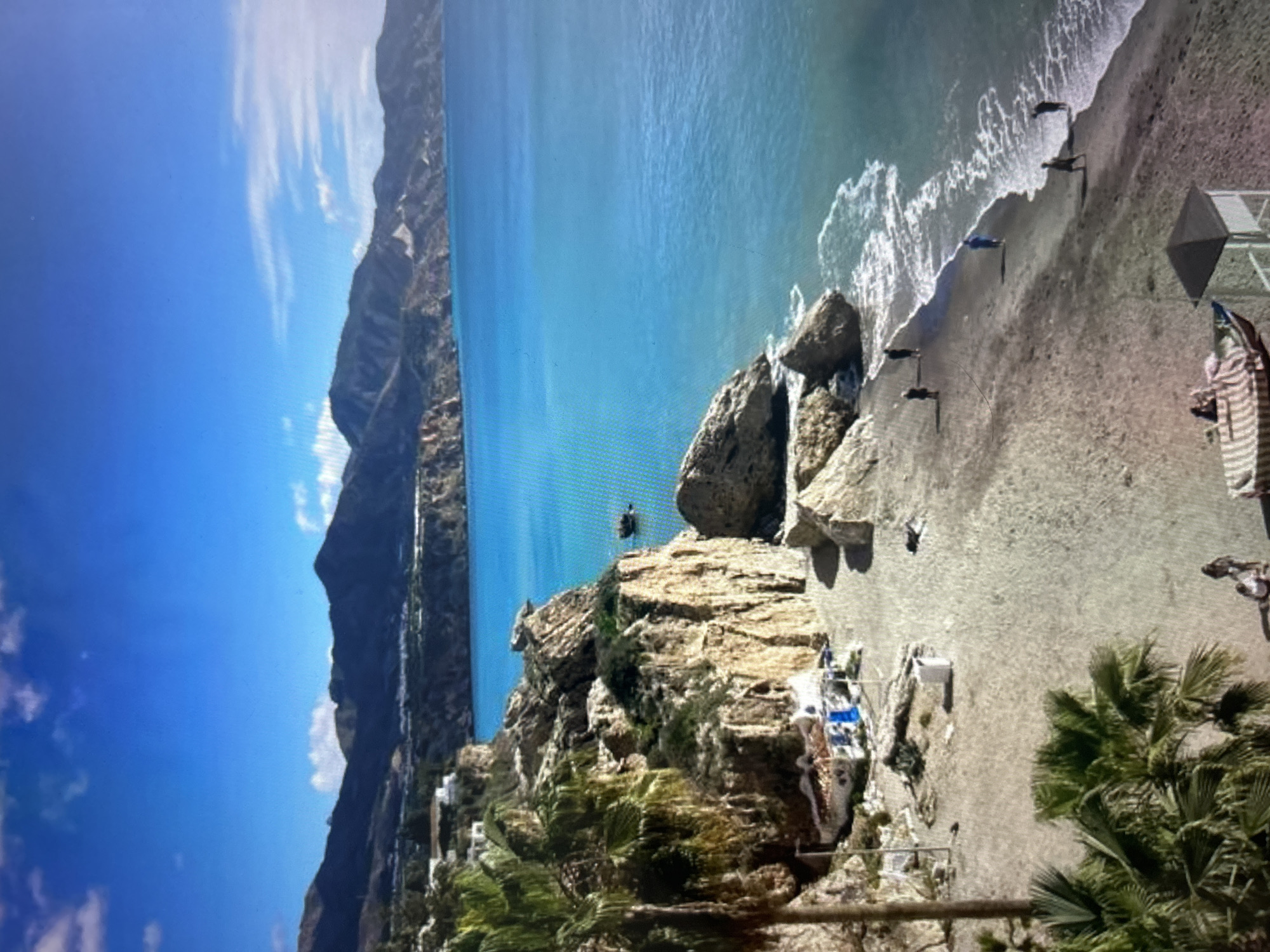 The image, viewed horizontally, captures a stunning coastal landscape from a high vantage point. A clear, vibrant blue sky with scattered white, wispy clouds dominates the upper portion. On the left, imposing, dark mountains or cliffs rise steeply, covered in patches of vegetation and dotted with scattered white buildings, possibly forming a village or town.
In the center, a calm expanse of bright turquoise-blue sea stretches towards the horizon. Gentle white waves break along a sandy beach located on the right side of the image. Numerous large, rugged rock formations are visible; some are integrated into the cliff face, while others stand as prominent outcrops near and in the water. A large rocky headland juts into the sea in the foreground, featuring some white and blue structures, likely beach facilities or small huts, nestled at its base.
Several small, dark figures, presumably people, are visible on the sandy shore and in the shallow water, suggesting they are swimming or wading. One solitary figure is perched high on the rocky headland, seemingly observing the scene. On the beach, near the water, a white and gray structure resembling a lifeguard station is present, along with a partially visible striped beach umbrella. Lush green palm trees and other foliage are seen in the immediate foreground, adding to the tropical or Mediterranean feel.
The scene depicts a sunny, clear day, likely midday given the bright illumination. The overall impression is that of a serene and picturesque holiday destination. No discernible text is visible in the image.