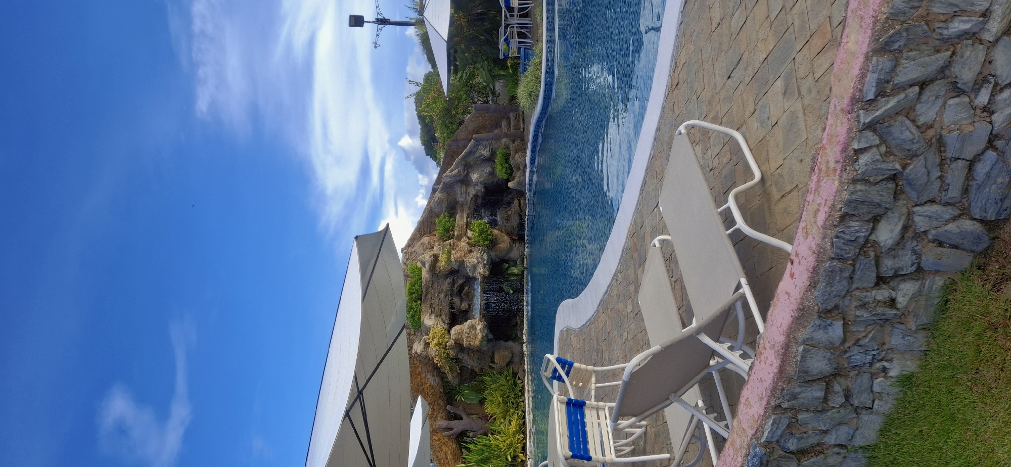 In Vallecito, Venezuela, this image captures a serene outdoor swimming pool area. The pool itself is a vibrant blue, with a cascading waterfall feature built into a rocky, landscaped mound at the far end. Lush green foliage surrounds the waterfall, adding to the tropical ambiance. Two white lounge chairs with light gray cushions are positioned on the paved stone patio in the foreground, suggesting a place for relaxation. In the background, large white umbrellas provide shade, and some tables and chairs are visible, hinting at an area for dining or socializing. The sky above is a bright blue, dotted with wispy white clouds, and a few small birds are visible in flight. The overall impression is one of a peaceful and inviting resort or private residence.