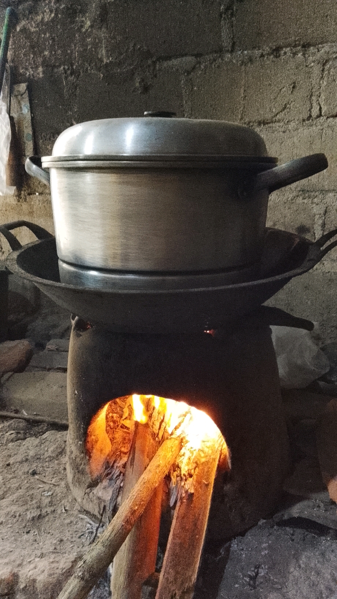 An outdoor scene in Way Melan, Indonesia, shows a clay stove with a fire burning inside. A large metal pot with a lid sits on top of a wok which is placed on the stove. The background consists of a rough, textured wall made of bricks. The ground is covered with dirt and debris.