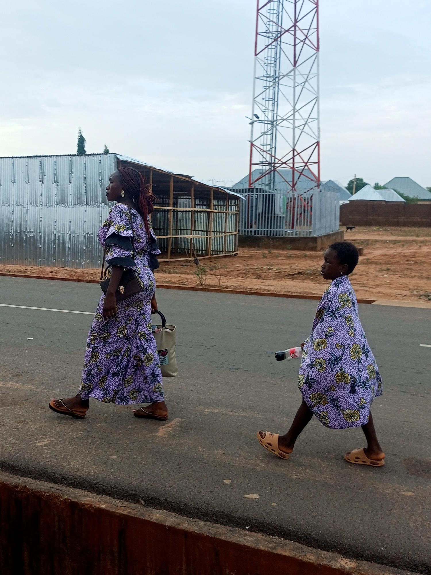 Two people, an adult and a child, are walking on a paved road in Tempure, Nigeria. The adult is on the left side of the frame, wearing a long, patterned, purple dress with a small black purse slung over their shoulder and carrying a light-colored tote bag. The child, to the right of the adult, is also wearing a patterned, purple dress that matches the adult's. The child is holding a plastic bottle. Both the adult and child are wearing sandals. A red and white telecommunications tower is visible in the background. Behind that is a mix of corrugated metal structures and residential buildings with sloping roofs. The sky is overcast. The ground off the side of the road is reddish-brown dirt. A short concrete wall lines the edge of the road in the foreground.