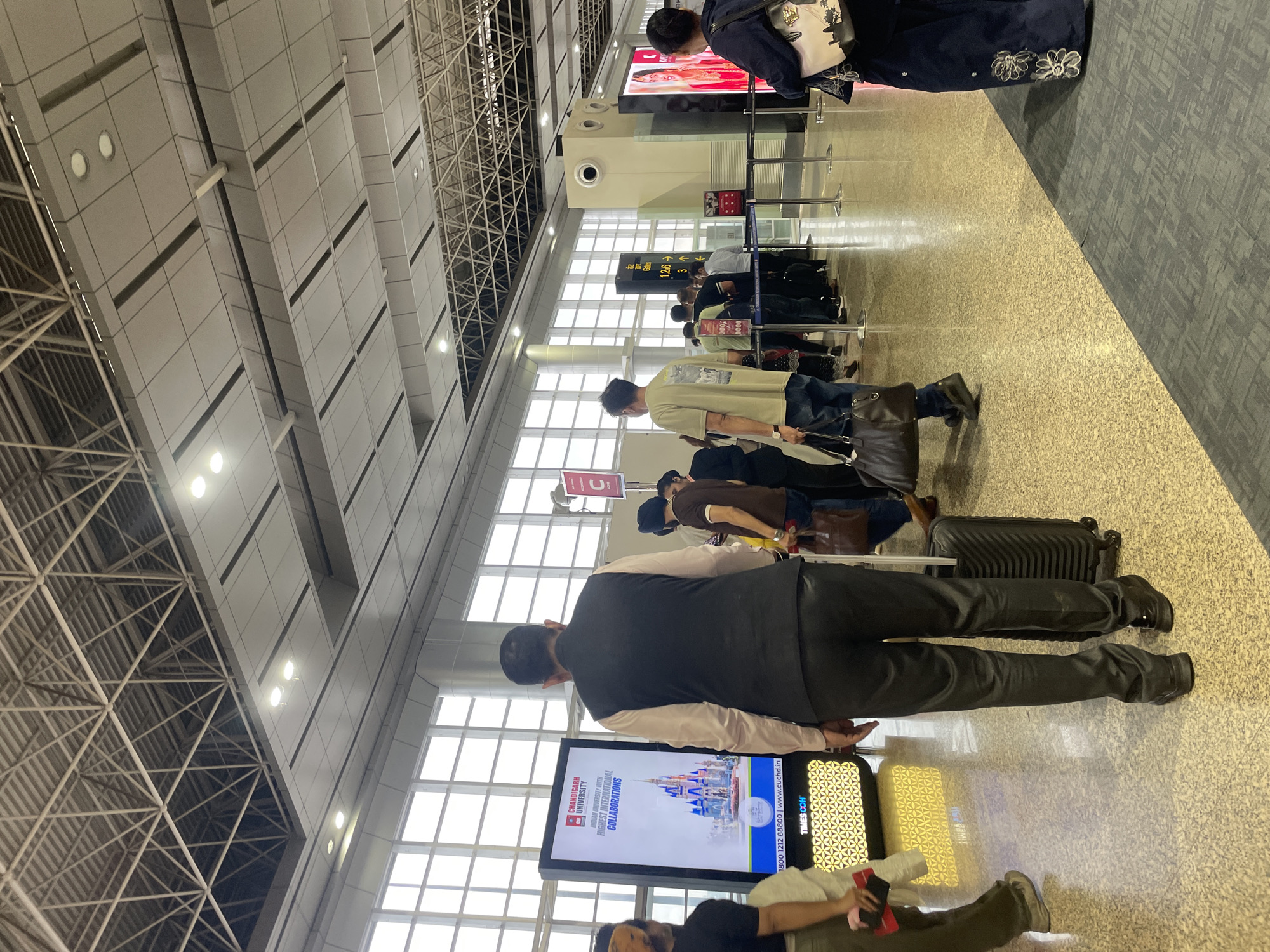 In the city of Raipur Khurd, India, an airport terminal shows a queue of people waiting. The terminal features a high ceiling with exposed metal trusses and modern lighting. Large windows line one side of the space, allowing natural light to enter. Several digital display screens are visible, one advertising Chandigarh University with an image of a castle. People are dressed in casual attire, with some carrying luggage and bags. The floor is a polished, speckled material. The overall scene depicts a typical airport environment with travelers in transit.