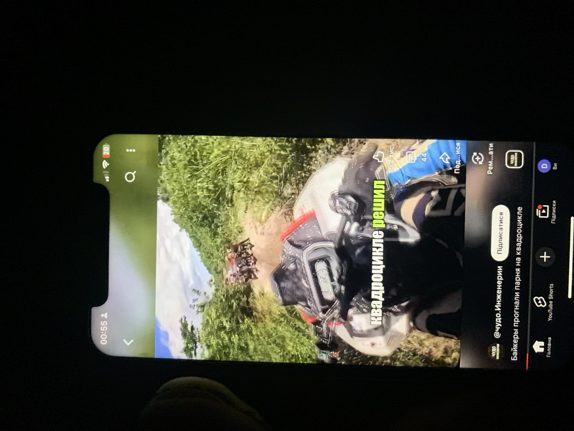 The image shows a close-up of a smartphone screen displaying a YouTube video. The video appears to be a first-person perspective of someone riding an ATV through a wooded, hilly area. The screen shows the video player controls, including a title overlay that reads "КВАДРОЦИКЛЕ РЕШИЛ" (which translates to "ATV decided" or "ATV made a decision"). Below the video, there are icons for likes, comments, shares, and subscriptions, along with channel information. The top of the screen shows the time "00:55" and other status icons. The background of the image is dark, indicating the photo was taken in low light conditions, likely at night. There is no indication of the city of Zhmerynka, Ukraine, in the image.