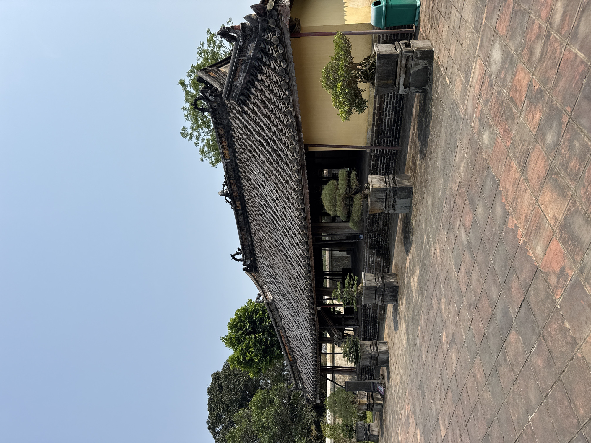 The image depicts a traditional Vietnamese building with a tiled roof in Kim Long Ward, Vietnam. The building features intricate architectural details, including ornate roof ornaments and supporting pillars. The tiled roof is composed of individual tiles arranged in a pattern. A tan building is in the background, with a green trash can on its right side. Trees surround the structure, adding greenery to the scene. The foreground consists of a paved walkway made of rectangular tiles. The sky is blue and clear.