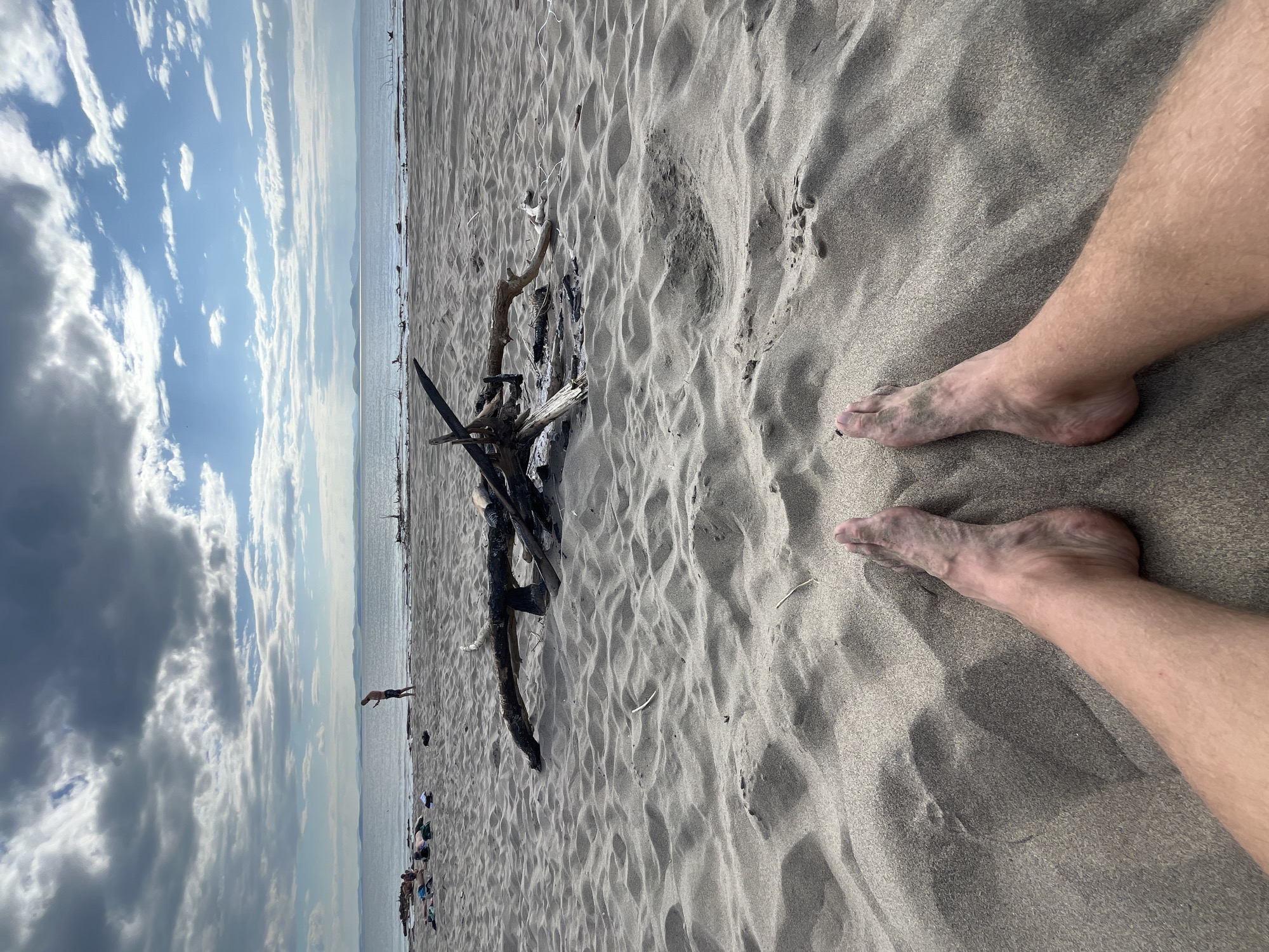 This image depicts a beach scene with a foreground dominated by sandy ground and a person's bare legs and feet. In the midground, there is a collection of driftwood, some of which appears to have been burned, suggesting a campfire site. Further in the background, the sea stretches out to the horizon, with hills visible in the distance under a cloudy sky with patches of blue. A few people are scattered on the beach, with one person standing near the water's edge. The scene is set on a beach, likely in the city of Krasnoturansk, Russia, given the context provided.