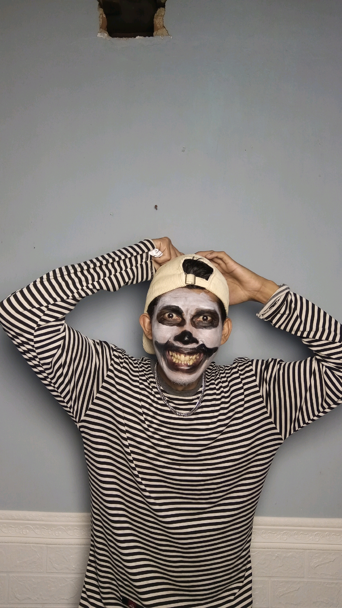 A person wearing a black and white striped long-sleeved shirt and a beige baseball cap is shown from the chest up, posing against a light blue wall. The person's face is painted with white makeup, resembling a skull, with black circles around the eyes and black paint accentuating the cheekbones and mouth. The person is smiling widely, revealing teeth. Their hands are placed on top of their head, near the cap. The wall behind the person has a rough, square hole near the top center. The bottom portion of the wall features a decorative white trim and a white brick-like texture. The image appears to be taken indoors. There is no visible indication of the location being in Kebumen, Indonesia.