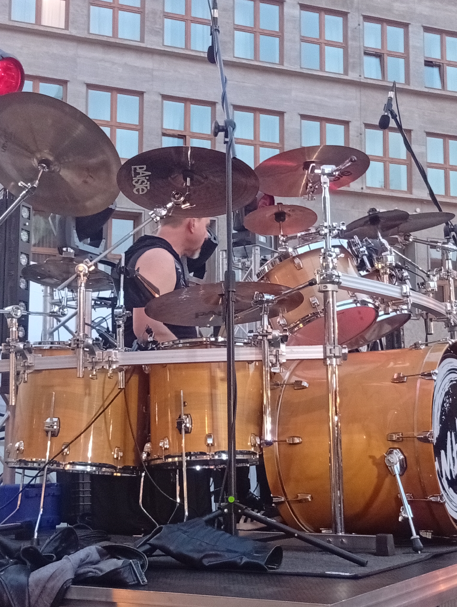 A male drummer, wearing a black sleeveless shirt, plays a drum set on a stage in front of a building with multiple windows. The drum set includes several cymbals and drums, with the wood grain of the drums visible. The building in the background appears to be made of concrete or a similar material and has a repeating pattern of windows with wooden frames. The scene suggests an outdoor performance, possibly a concert, given the stage setup and the presence of musical equipment. The lighting suggests it is daytime, but the sky is not visible. The location is the Wrocław Congress Center, Poland.