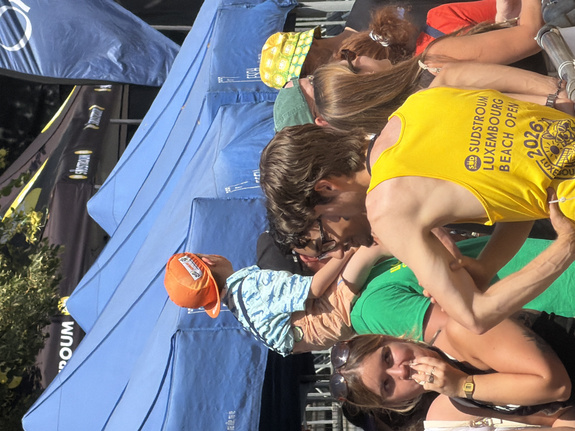 The image depicts an outdoor event under blue tents, likely in Esch-sur-Alzette, Luxembourg, given the text "SUDSTROUM LUXEMBOURG BEACH OPEN 2026" visible on a yellow tank top. In the foreground, a young child wearing an orange cap is being carried on someone's shoulders. The child is dressed in a light blue shirt with a dinosaur pattern and orange shorts. Several other individuals are visible in the mid-ground and background, some looking towards the camera, others engaged in conversation or observing the event. The environment appears to be a public gathering with branding banners in the background.