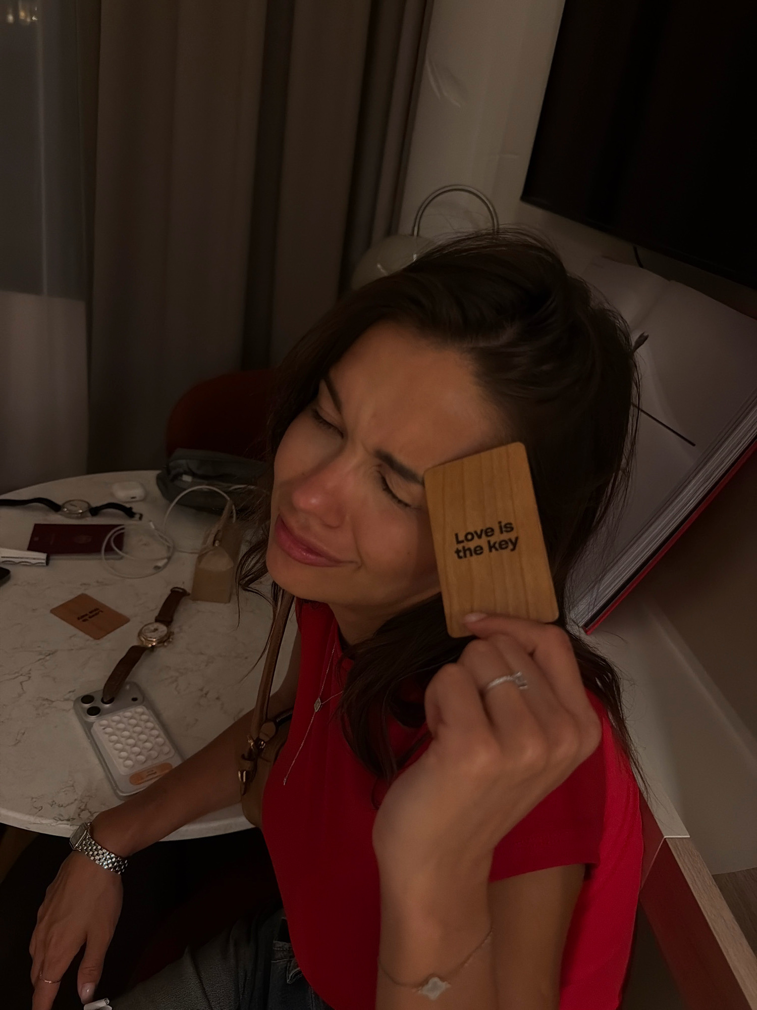 A woman with dark hair, wearing a red top and a ring on her right ring finger, is holding a wooden card with the text "Love is the key" printed on it. She is looking down with her eyes closed. On the table in front of her, there are two watches, a passport, a phone case with many white circles, and a brown paper bag. The background consists of dark curtains and what appears to be a television screen. The scene is indoors, likely in a hotel room, given the presence of a passport and room key card. The lighting suggests it is evening or the room is dimly lit. There is no indication of the city of Amsterdam, Netherlands within the image.