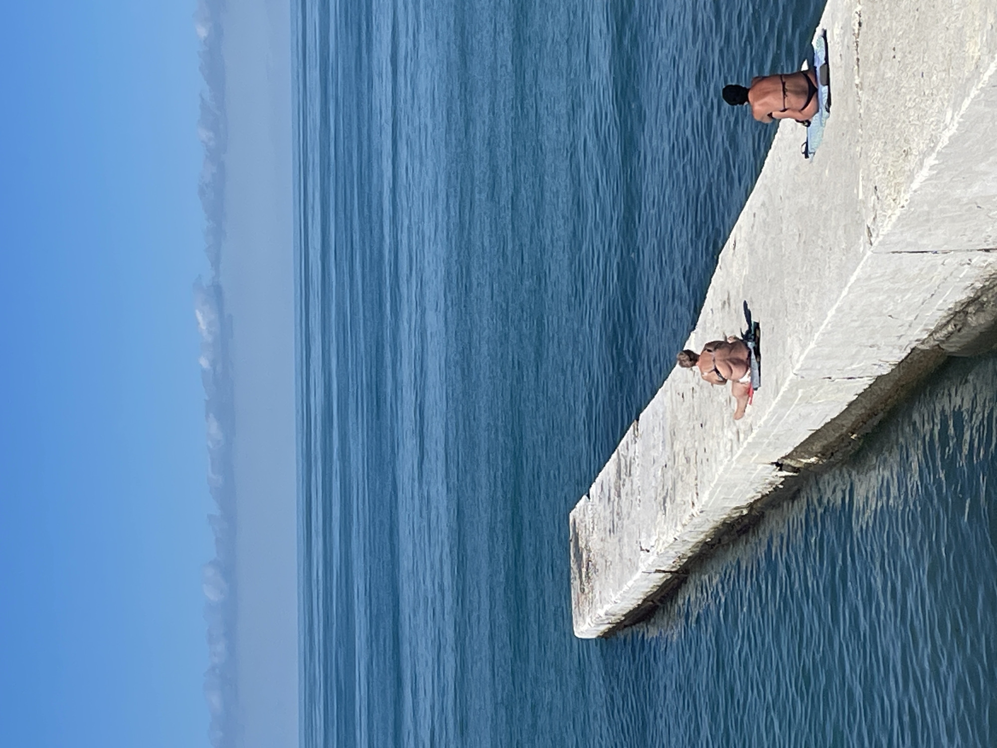 Two individuals are sunbathing on a concrete pier extending into the blue sea. The pier is long and rectangular, with some wear and tear visible on its surface. The water is calm, with gentle ripples. In the background, a hazy horizon line separates the sea from a light blue sky with a few wispy clouds. The scene appears to be a typical summer day at a seaside location. The image does not contain any explicit visual cues to identify it as being in Sochi, Russia.