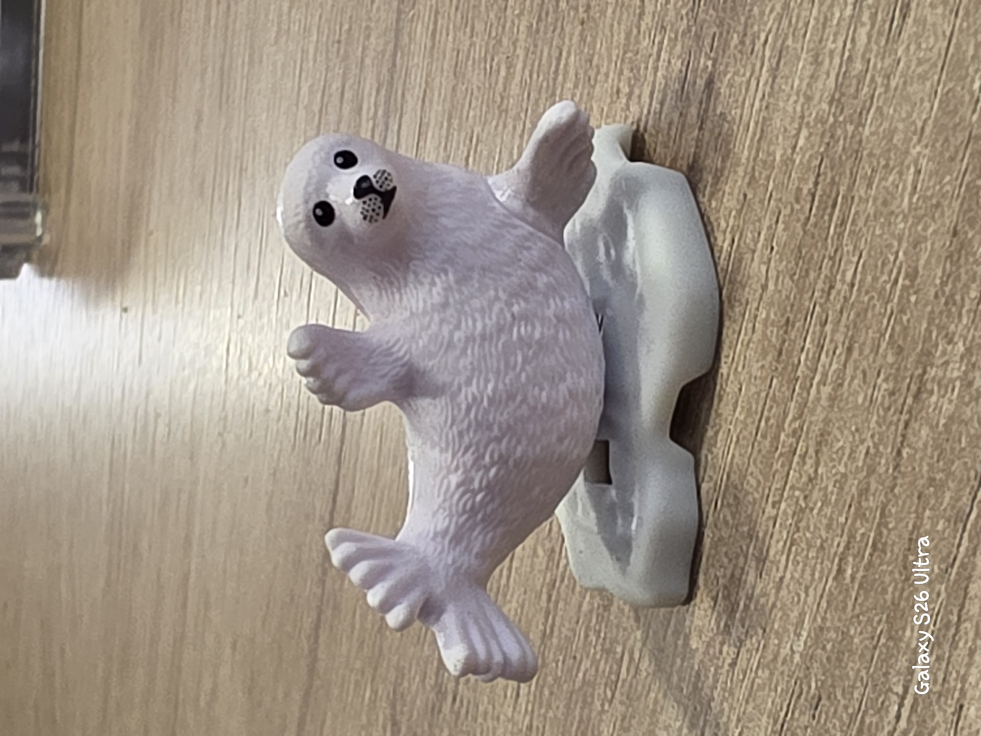 A light purple toy seal is positioned on a light gray, iceberg-shaped platform, which rests on a wooden surface. The seal is lying on its back with its flippers raised, facing left. Its eyes are black circles, and it has a black, speckled muzzle. The wooden surface appears to be a table or floor with a light brown, grain pattern. The background is blurred and shows a dark, reflective surface. In the lower left corner, the text "Galaxy S26 Ultra" is visible, indicating the device used to capture the image. This image does not depict any specific location in Plovdiv, Bulgaria.