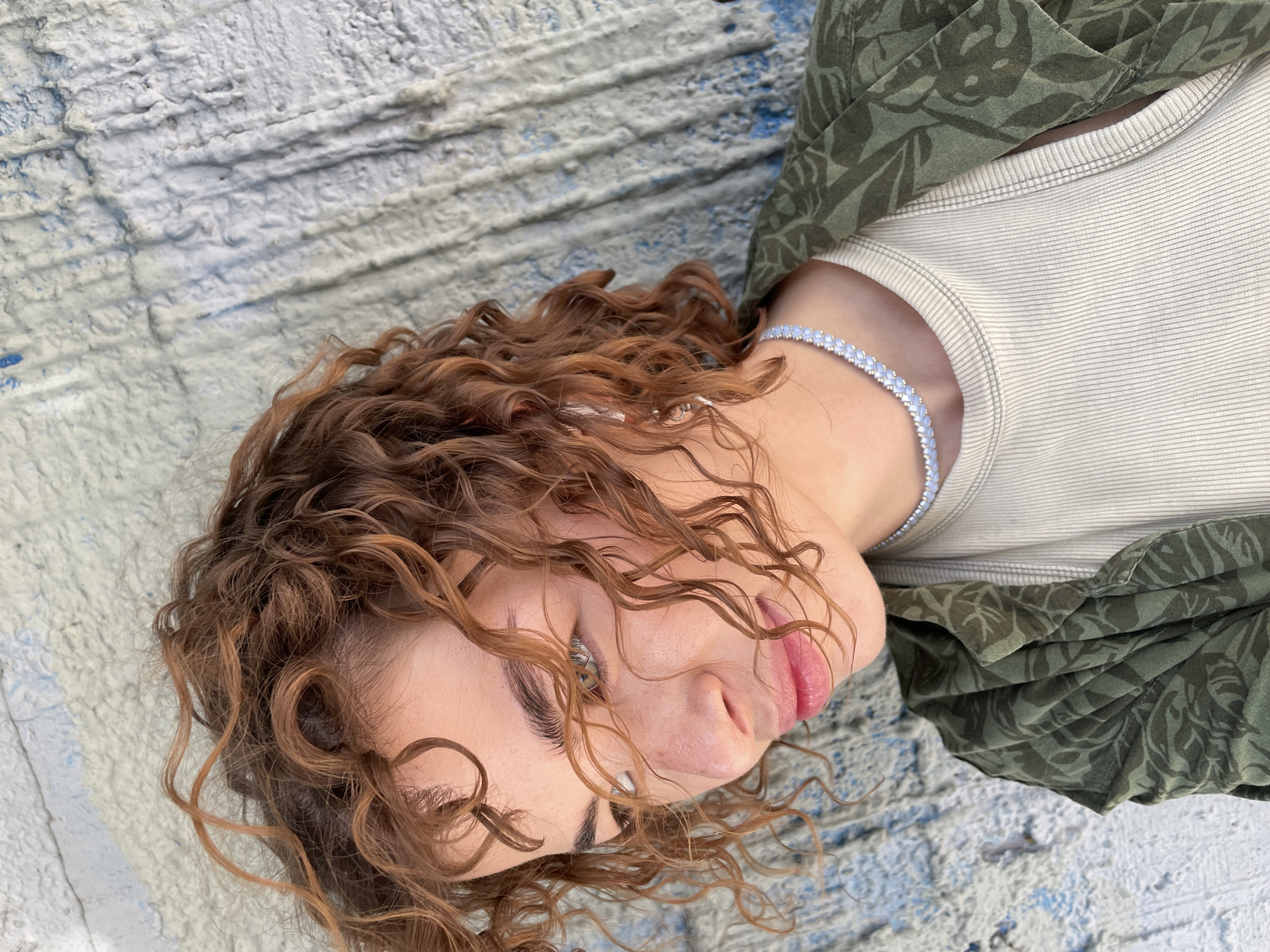 A woman lies on a textured surface painted with white and blue paint. She has curly brown hair and is wearing a light-colored tank top, a green patterned shirt and a beaded necklace. The location is not specified in the image.