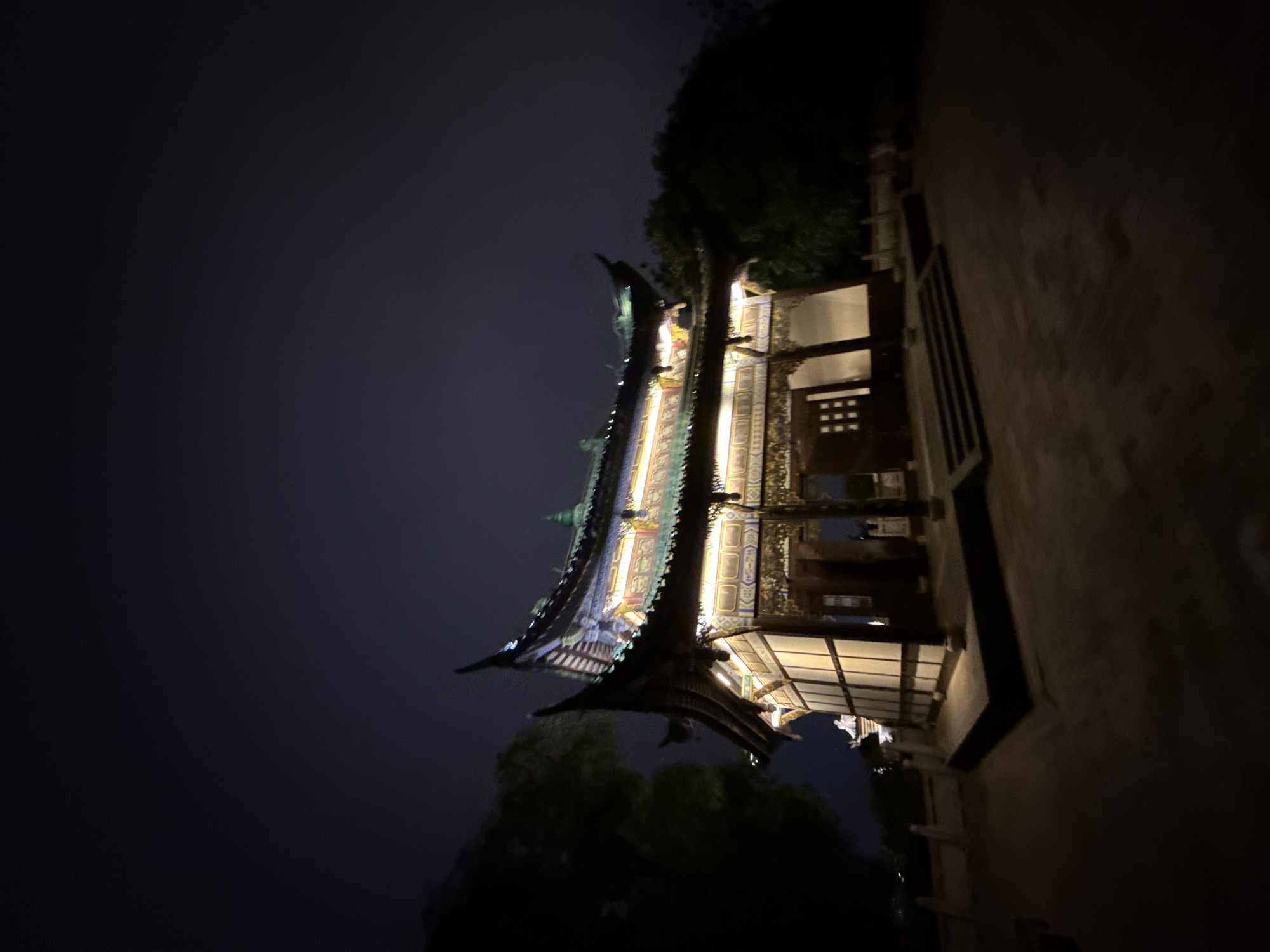 A nighttime shot captures an ornate Chinese-style building against a dark sky, likely in Liangjiang, China. The building features tiered, upturned eaves adorned with intricate decorations and illuminated by internal lighting. Trees are partially visible at the building's base. A walkway or wall borders the structure.