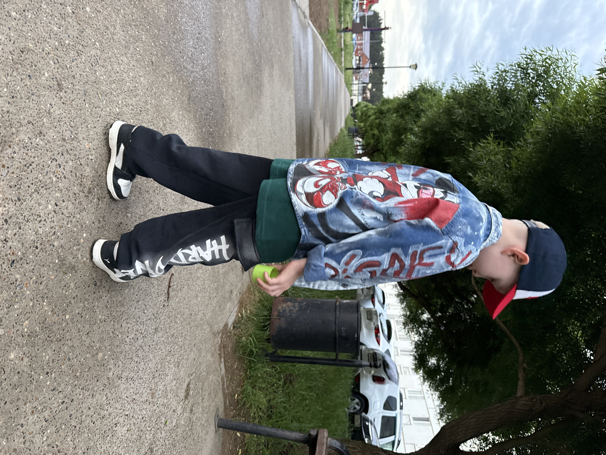 A child stands outdoors on a paved walkway, likely in a park setting, near a black metal trash receptacle. The location is potentially Chaikovsky, Russia. The child wears black pants with white lettering, black and white sneakers, a green shirt, and a denim jacket with a graphic design. They are also wearing a dark baseball cap with a red brim. In their hand, they hold a small, green object. Behind them, there is a grassy area, trees, parked white cars, and buildings. A pathway extends further into the background. The sky is overcast.