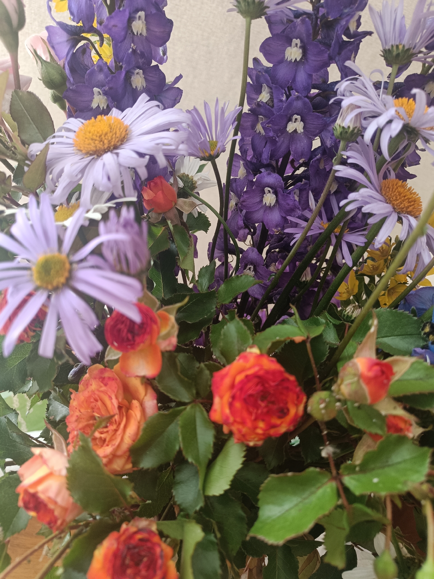 This media file features a close-up, overhead shot of a vibrant floral arrangement. The bouquet is densely packed with a variety of flowers in shades of purple, lavender, orange, and yellow, interspersed with lush green foliage. Dominating the composition are tall, spiky purple delphiniums and large, daisy-like flowers with pale lavender petals and bright yellow centers. Smaller, delicate lavender asters with thin petals are also visible, along with clustered sprays of small orange and red roses, some still in bud. Tiny yellow flowers add pops of contrasting color to the background.
The lighting suggests an indoor setting or an overcast day, as there are no strong shadows, and the overall tone is soft. The focus is primarily on the flowers, with the background appearing slightly blurred. The arrangement is presented in a way that emphasizes its texture and color variety. There are no people or discernible objects other than the flowers and their stems and leaves. The scene evokes a sense of natural beauty and perhaps a celebration or a gift. No text is visible in the image. The location context provided is Первомайское, Russia, which, while not visually evident in the image itself, suggests a possible origin or context for the floral arrangement.