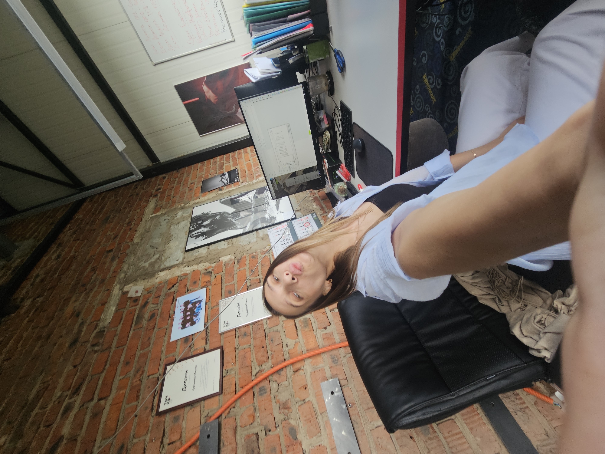 A young woman is taking a selfie in a brick-walled office. She is sitting in a black leather office chair and wearing a light blue open shirt over a black tank top and white pants. She is puckering her lips towards the camera. On the brick wall behind her are two framed certificates and a framed group photograph. A black computer monitor with a spreadsheet on it is visible on a desk to her right, along with a keyboard, mouse, a glass of water, and a pair of scissors. The office appears to be well-lit with overhead fluorescent lighting and natural light from a window or opening not visible in the frame. The floor is carpeted with a circular pattern. There is no identifiable information to place the image in the city of Chaikovsky, Russia.